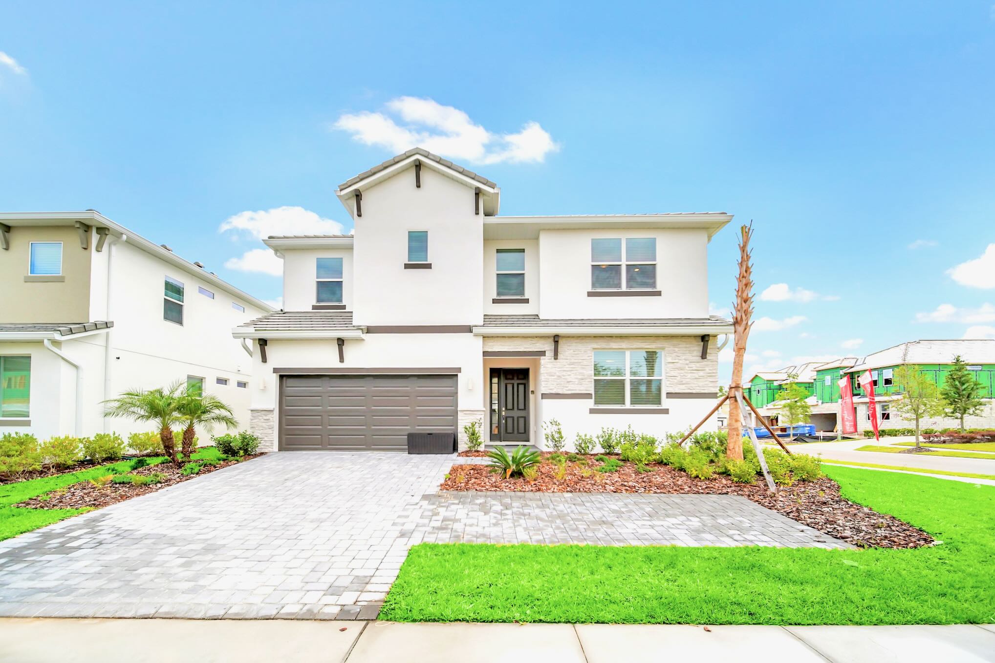 A modern two-story house with white stucco and stone accents features a paved driveway leading to a two-car garage, a dark front door, and well-maintained green lawns and landscaping.