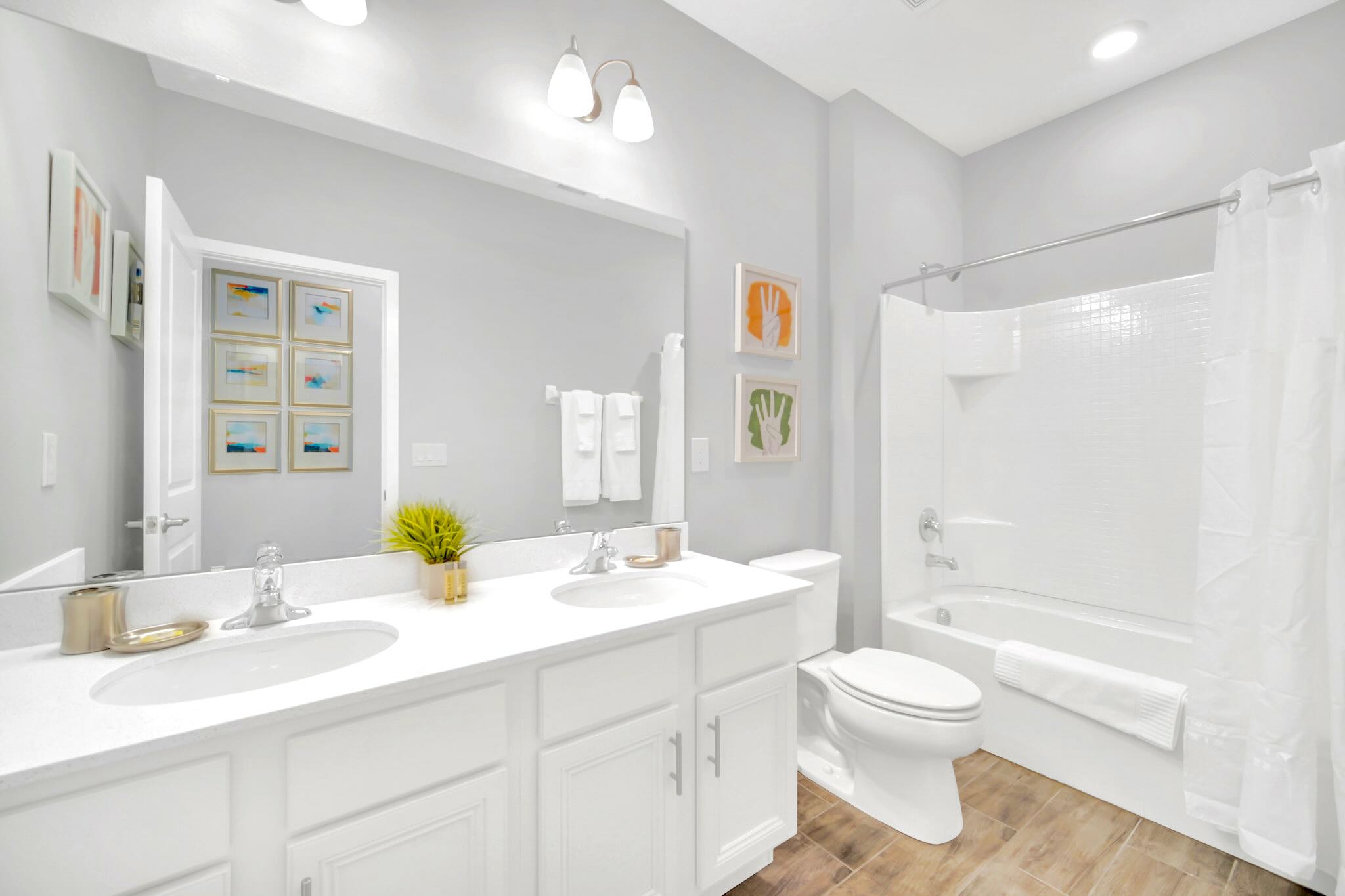 Bright bathroom with light gray walls, a white double vanity featuring two sinks, chrome faucets, and a large mirror. A white toilet and a shower/tub combo with a white curtain are visible on the right.