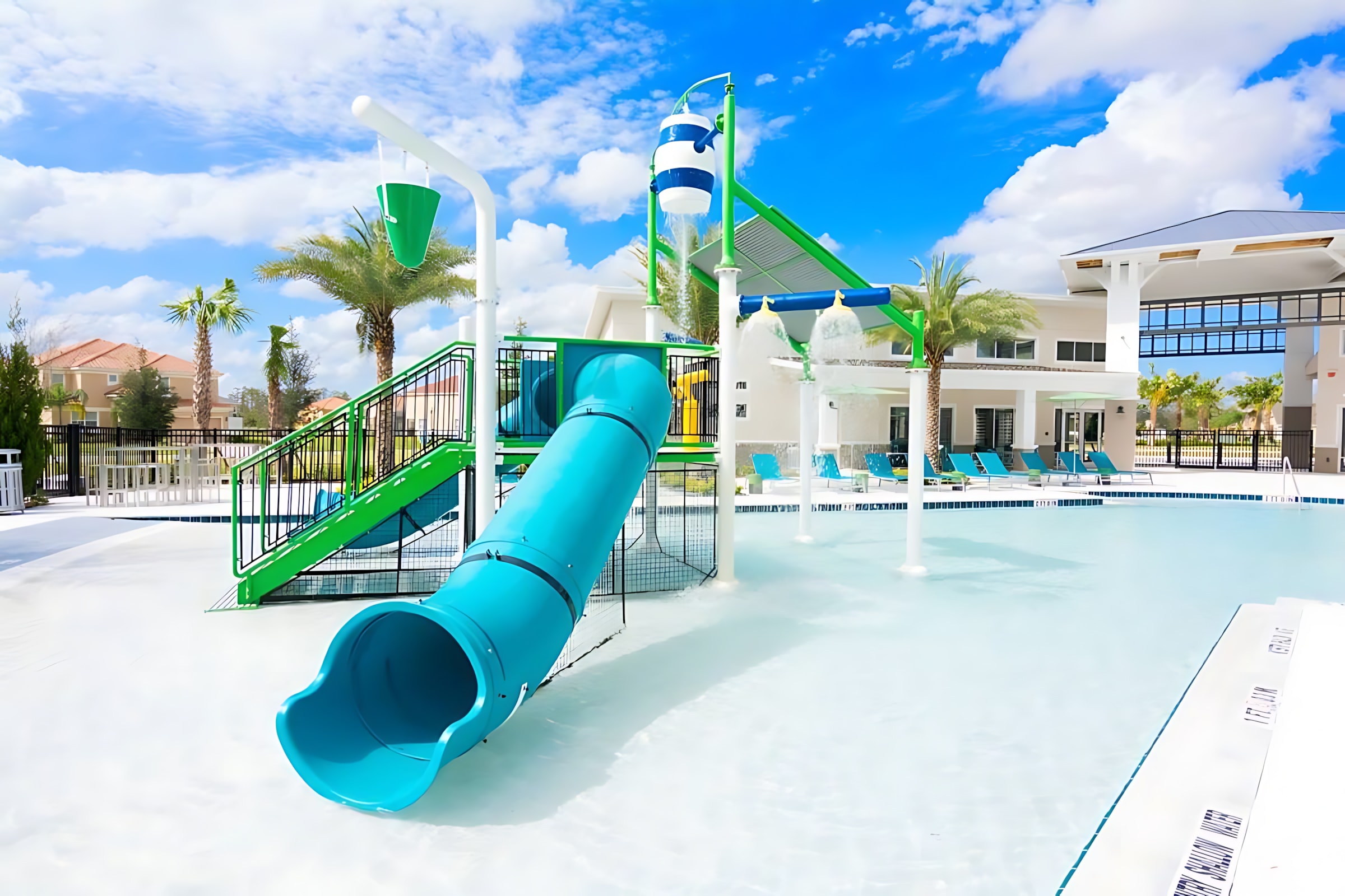 A vibrant communal pool area features a shallow entry with a blue and green water slide, a large dumping bucket, and various water sprayers, all under a bright blue sky with white clouds. Palm trees and a modern building with white walls and dark accents are visible in the background.