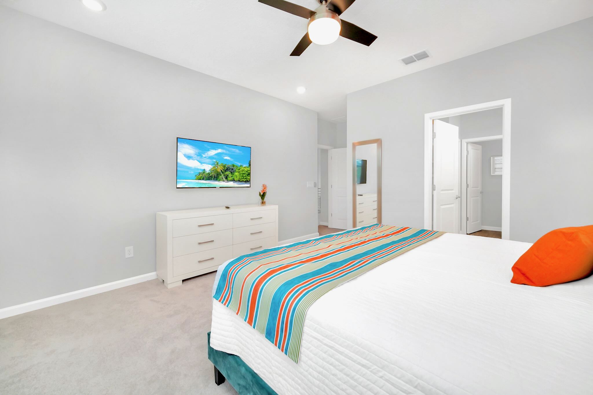 A bright bedroom with light gray walls, a white dresser with a TV displaying a beach scene, and a bed with a white comforter and a colorful striped throw. A dark ceiling fan with a light is visible overhead.