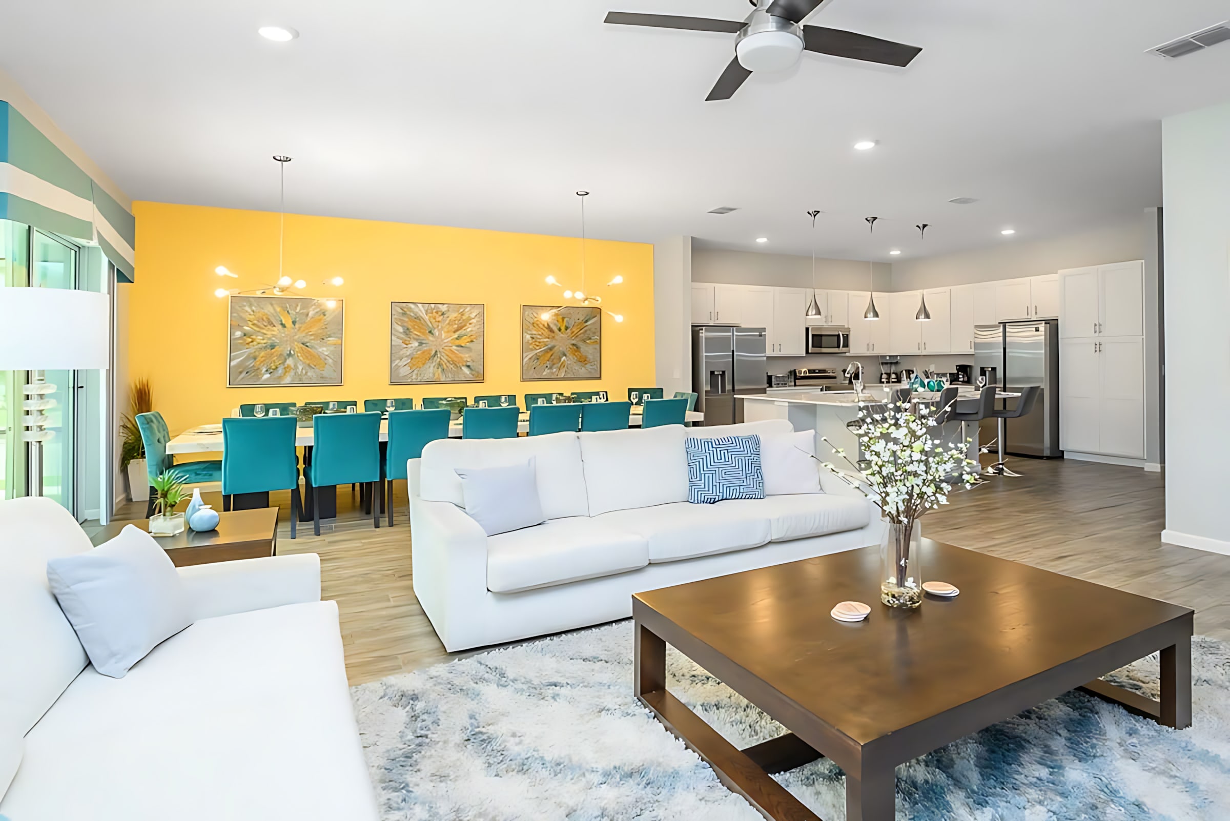 An expansive open-concept living space combines a bright living room with two white sofas and a coffee table, flowing into a modern kitchen with white cabinetry, stainless steel appliances, and a large island.
