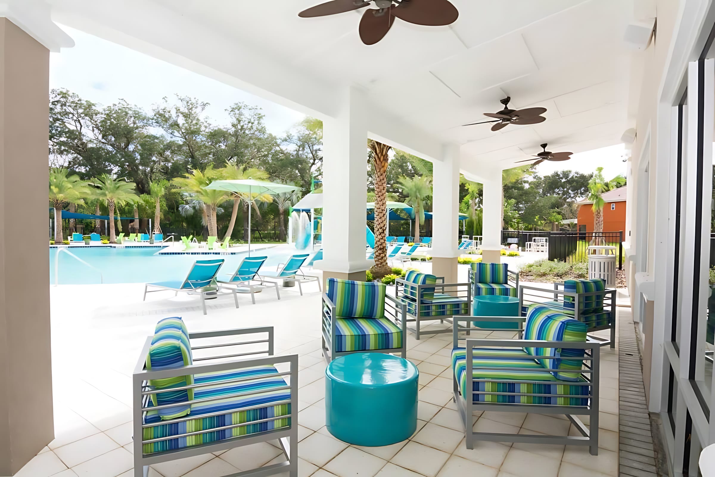 A covered outdoor patio area features several comfortable armchairs with blue and green striped cushions and a round turquoise coffee table. Ceiling fans are mounted above, and the communal pool area with lounge chairs and palm trees is visible in the background.
