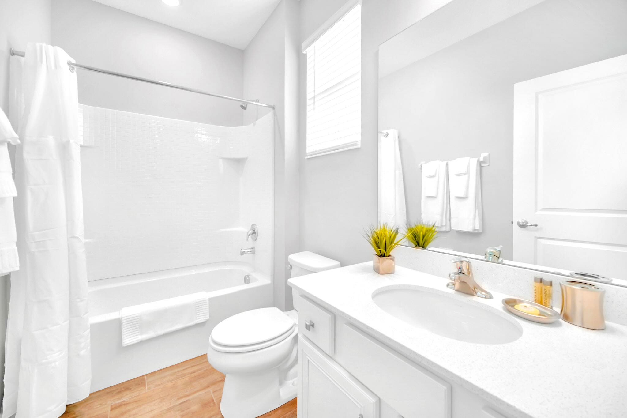 A bright, all-white bathroom featuring a single vanity with a white countertop, a large mirror, and a toilet. It includes a white bathtub with a shower curtain and light wood-look tile flooring, with a small window providing natural light.