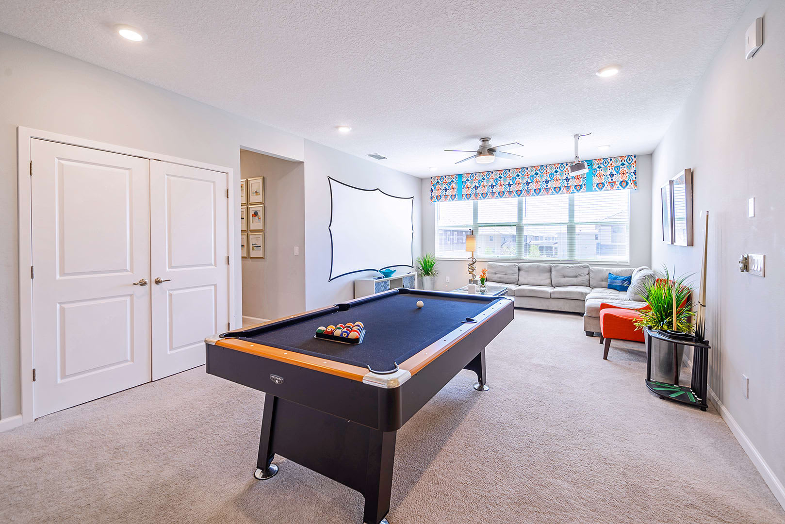 Spacious game room featuring a black pool table with cues, alongside a comfortable grey sectional sofa and a projector screen for entertainment. Double white doors are visible on the left, and large windows brighten the space.