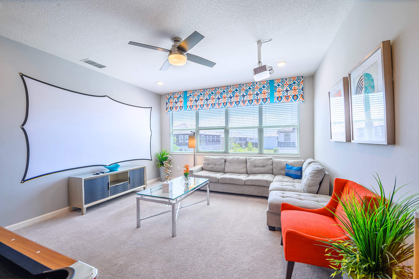 A cozy media room features a large projector screen, a comfortable L-shaped sectional sofa, an orange accent chair, and a glass coffee table, all under a ceiling fan and recessed lighting.