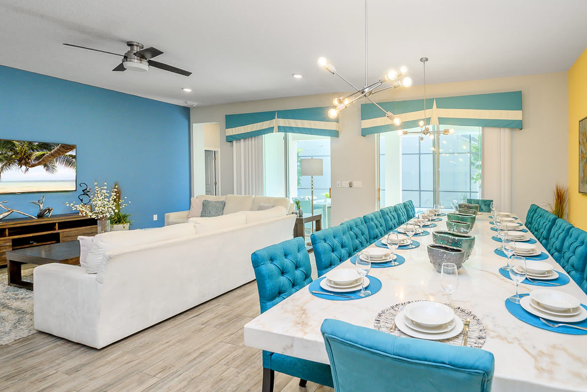 Long dining table with teal upholstered chairs, set with place settings, extending towards a bright living area with white sofas and a blue accent wall. Large windows with decorative valances offer views to the outside.