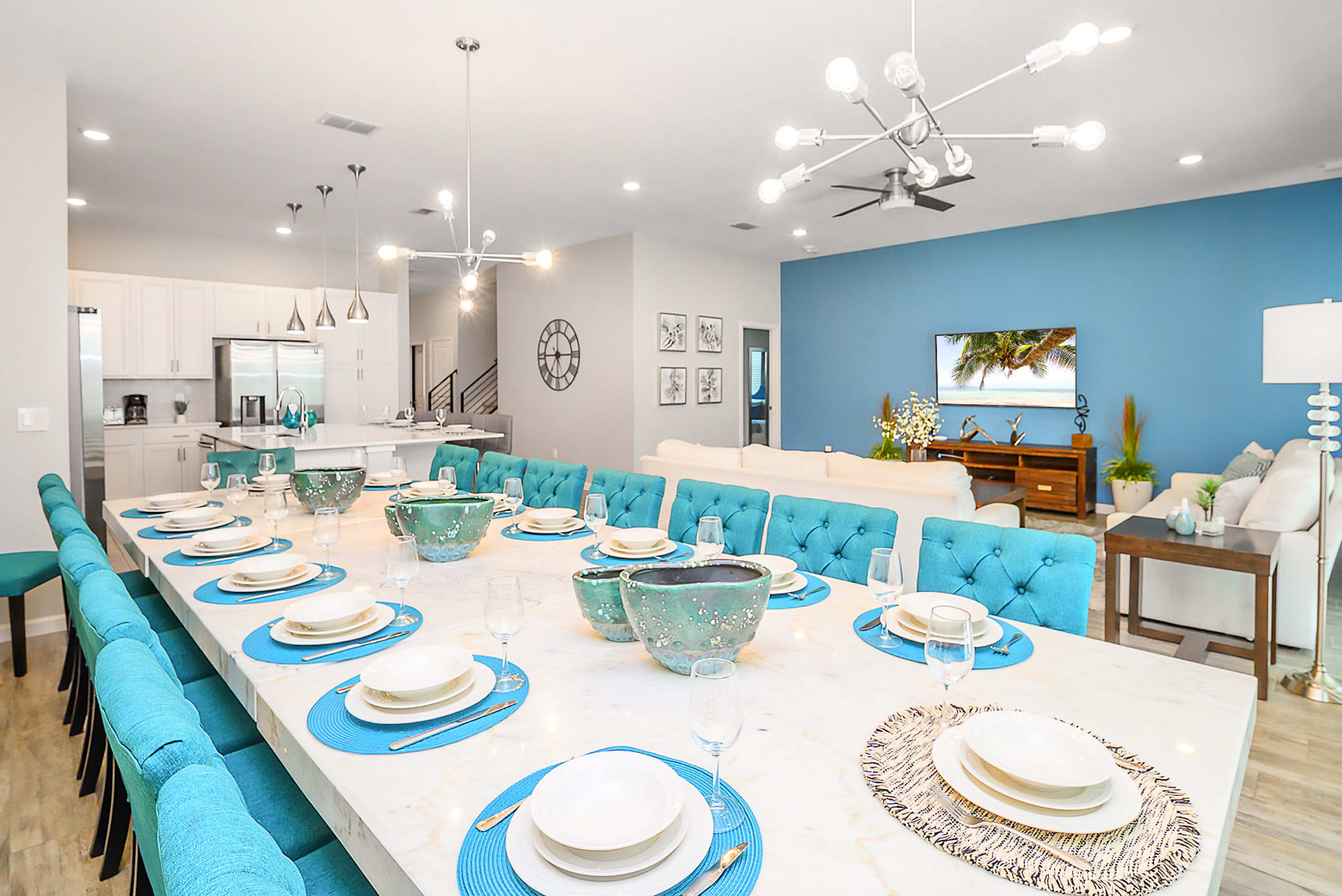 Expansive dining room featuring a long white marble-top table set with place settings and teal chairs, illuminated by modern linear chandeliers. The adjacent living room with a blue accent wall and a modern kitchen are visible in the background.