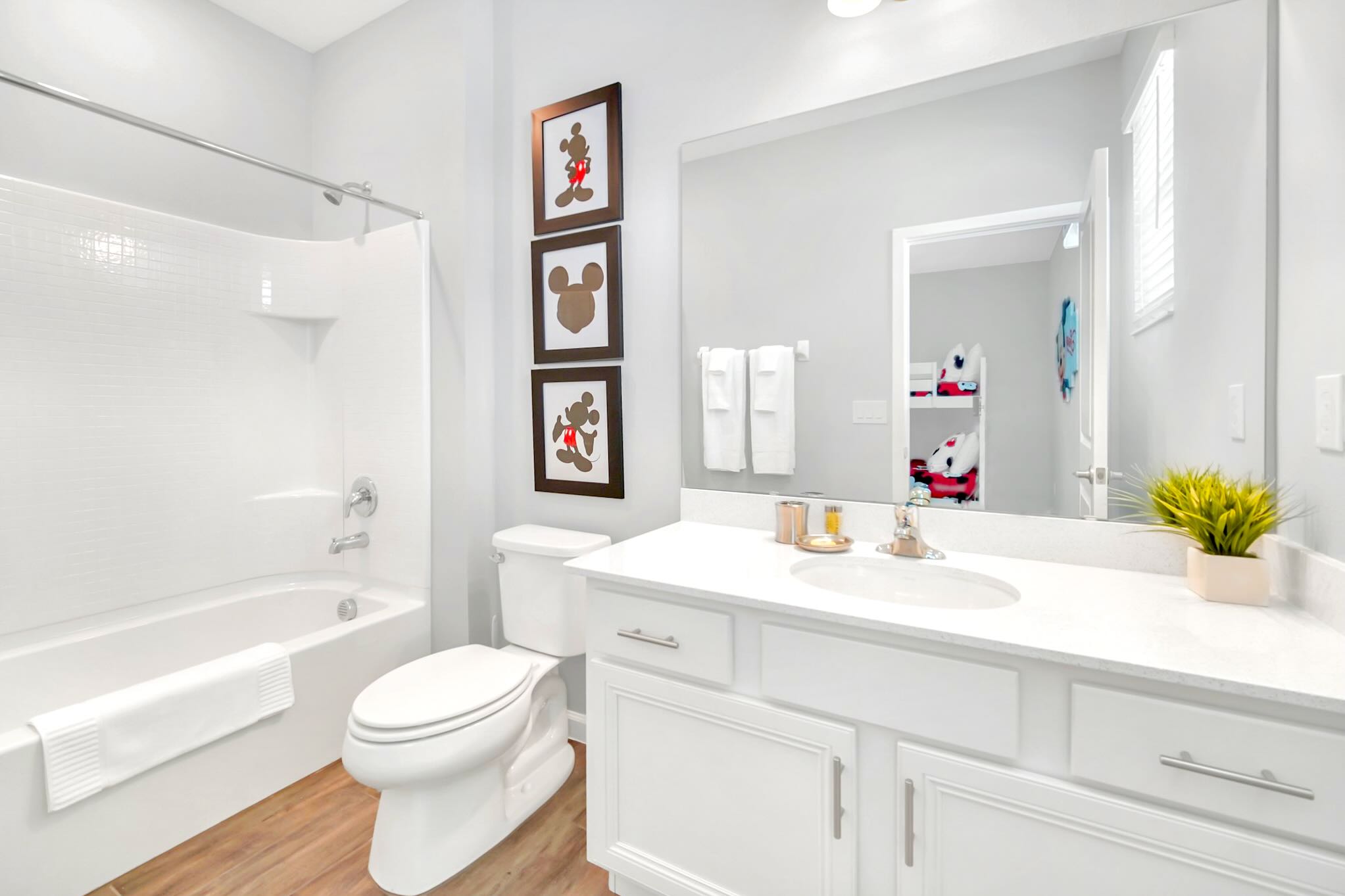 Modern bathroom with a white single vanity, chrome faucet, and a large mirror reflecting a bunk bed. It features a white toilet, a shower/tub combo, and Mickey Mouse framed art on the wall.