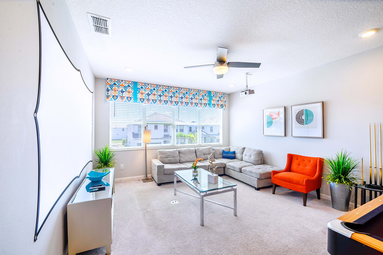 Bright media room featuring a large grey sectional sofa, a glass coffee table, and a projector screen on the wall. Large windows with colorful valances provide natural light, and an orange accent chair adds a pop of color.