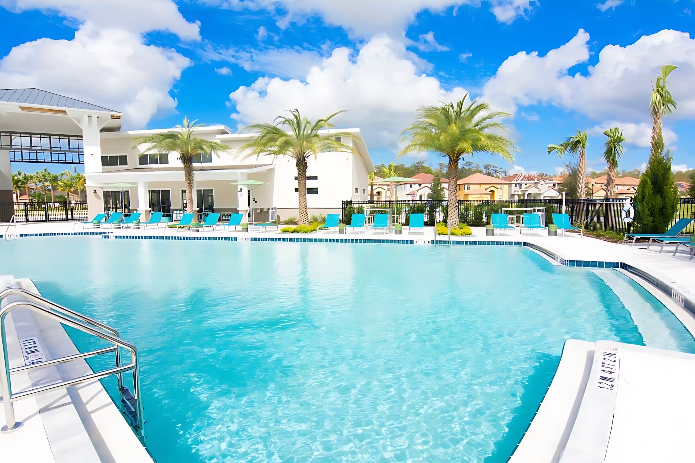 Large, rectangular communal swimming pool with clear blue water, surrounded by numerous blue lounge chairs and palm trees. The modern clubhouse building is visible in the distance under a bright sky.