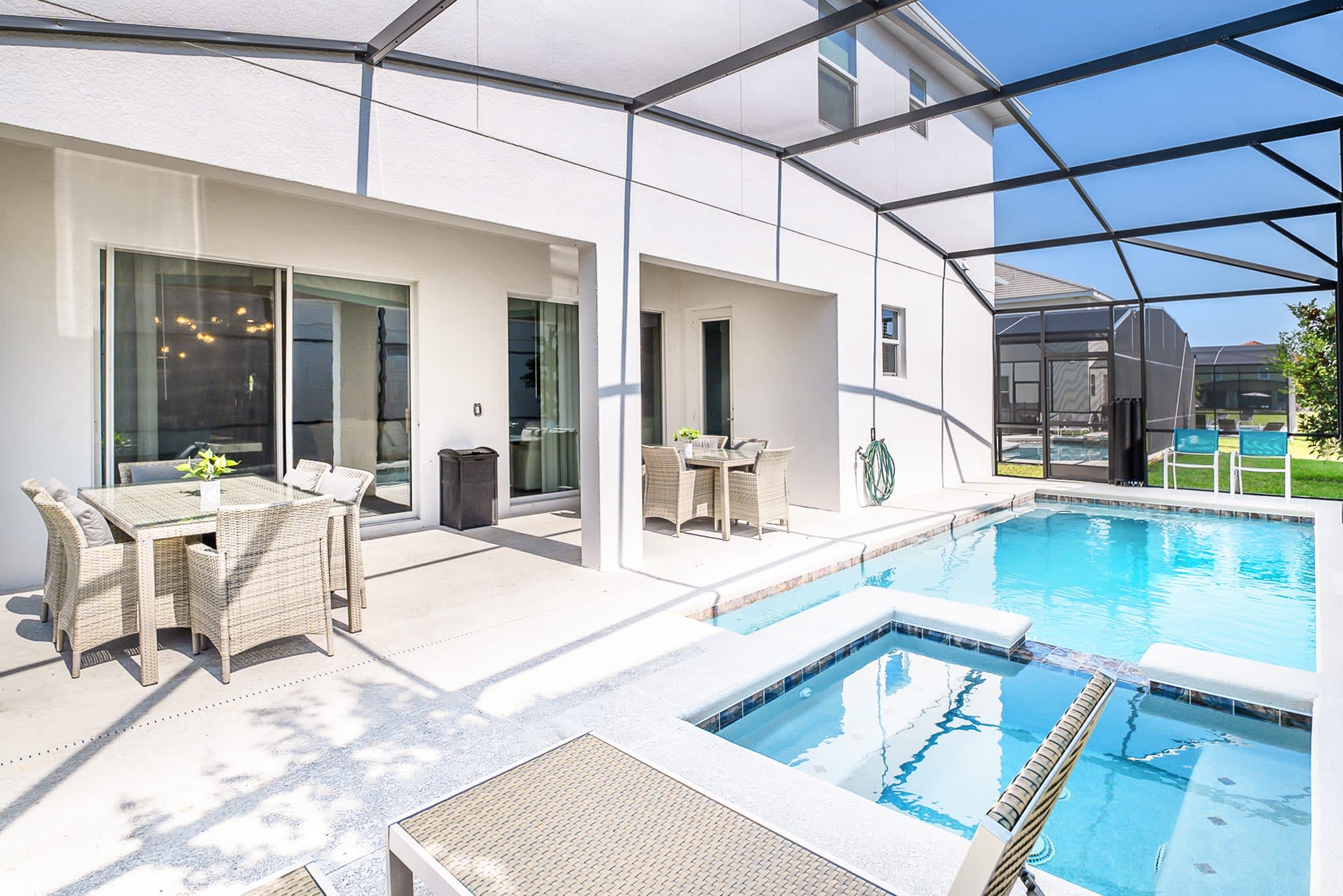 A bright outdoor patio features a large wicker dining table with a glass top and cushioned chairs, alongside a private swimming pool and integrated hot tub, all enclosed within a screened lanai.