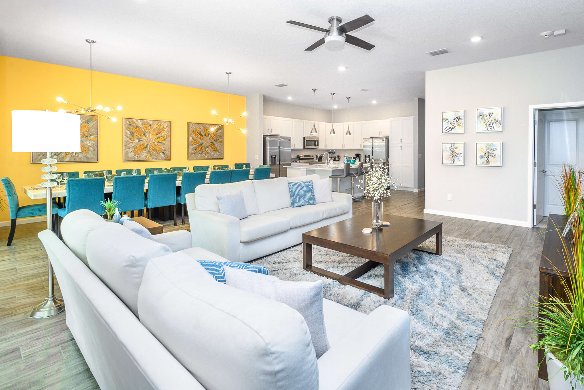Bright open-concept living space featuring white sofas, a dark wood coffee table on a blue and white shag rug, and a dining area with a vibrant yellow accent wall and teal chairs. A modern kitchen with white cabinets and stainless steel appliances is visible in the background.