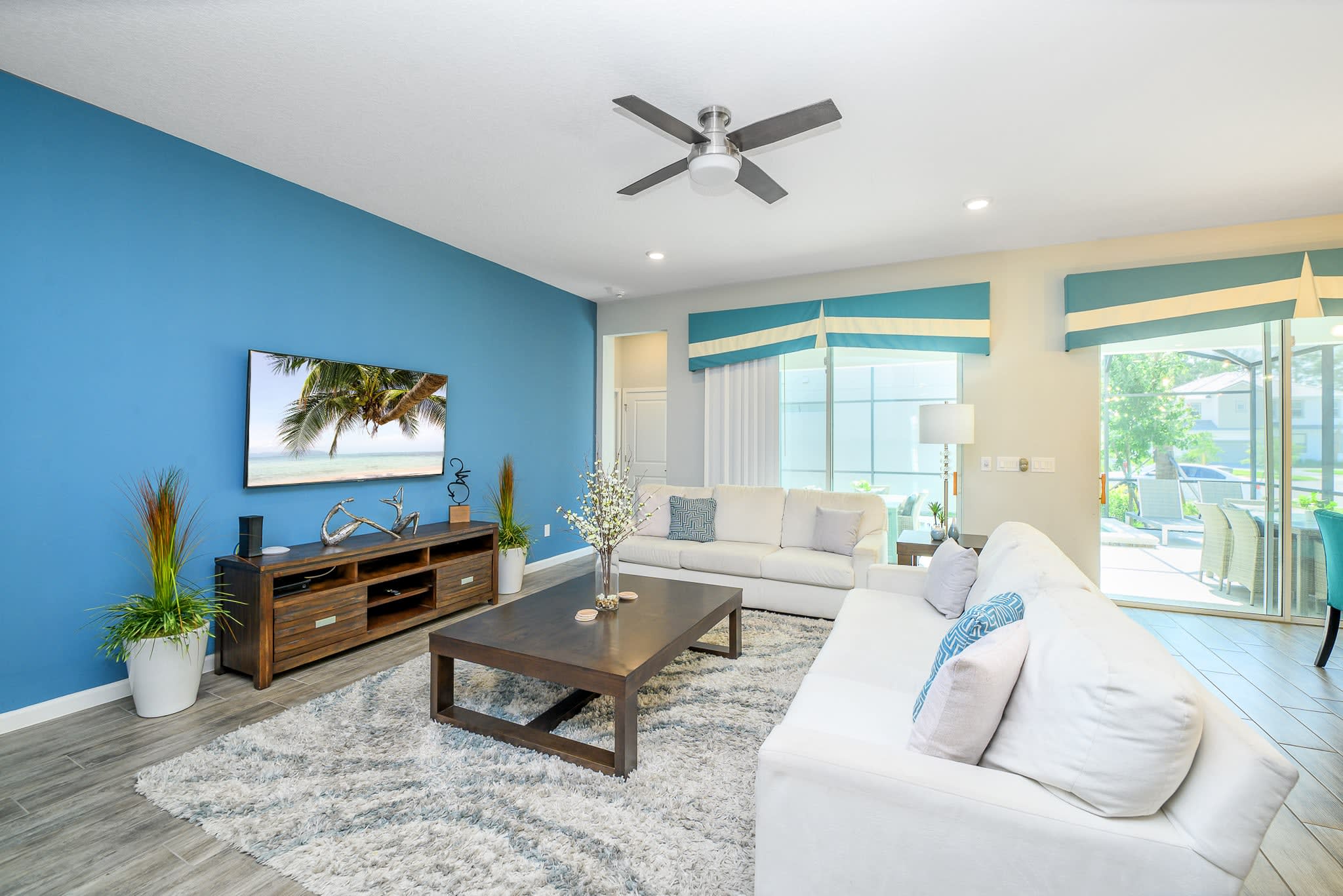 Cozy living room with a striking blue accent wall featuring a large wall-mounted TV above a wooden media console. Two white sofas face each other, centered around a dark wood coffee table on a plush grey and white rug.