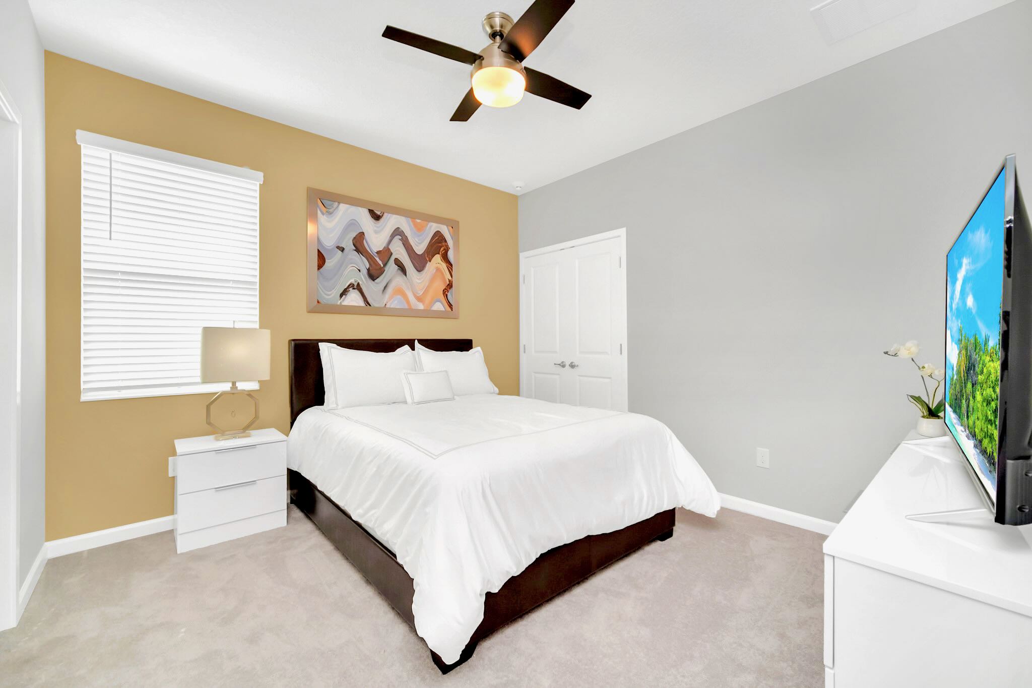 This bedroom features a queen-sized bed with a dark brown upholstered frame and crisp white bedding, set against a warm beige accent wall. The room includes a ceiling fan, a white nightstand with a lamp, and a TV on a matching dresser.
