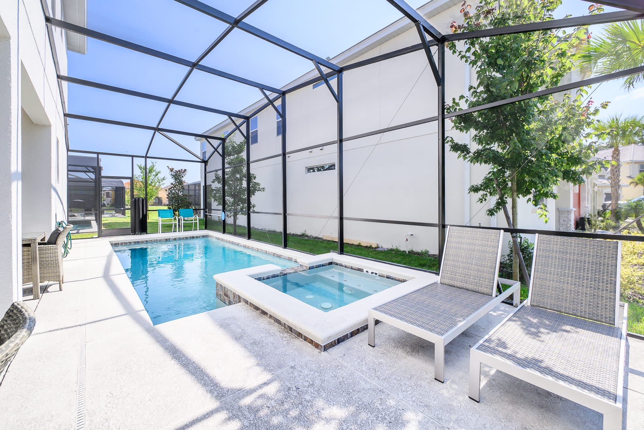 A private screened pool area features a rectangular swimming pool and an adjacent hot tub, surrounded by a light-colored patio deck and two lounge chairs, with lush greenery outside the enclosure.