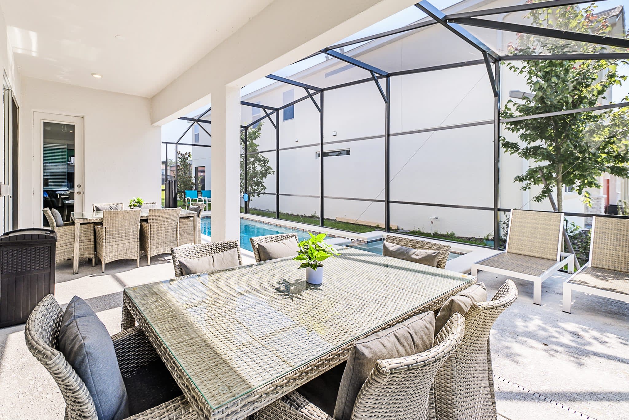A covered outdoor patio area under a screened enclosure features a large wicker dining table with a glass top and cushioned chairs, offering a comfortable space for al fresco meals.