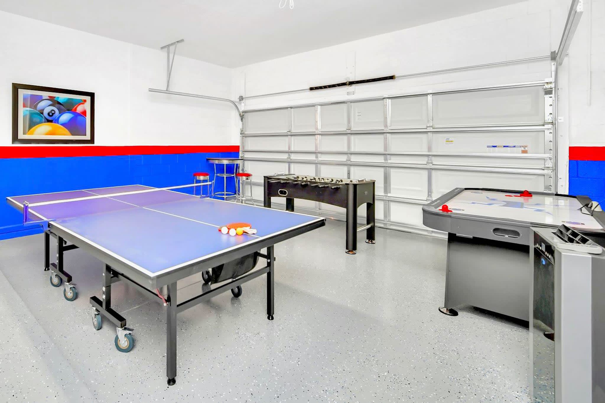 A spacious game room, housed in a garage, features a blue ping pong table, a foosball table, and an air hockey table, with a large garage door visible in the background.