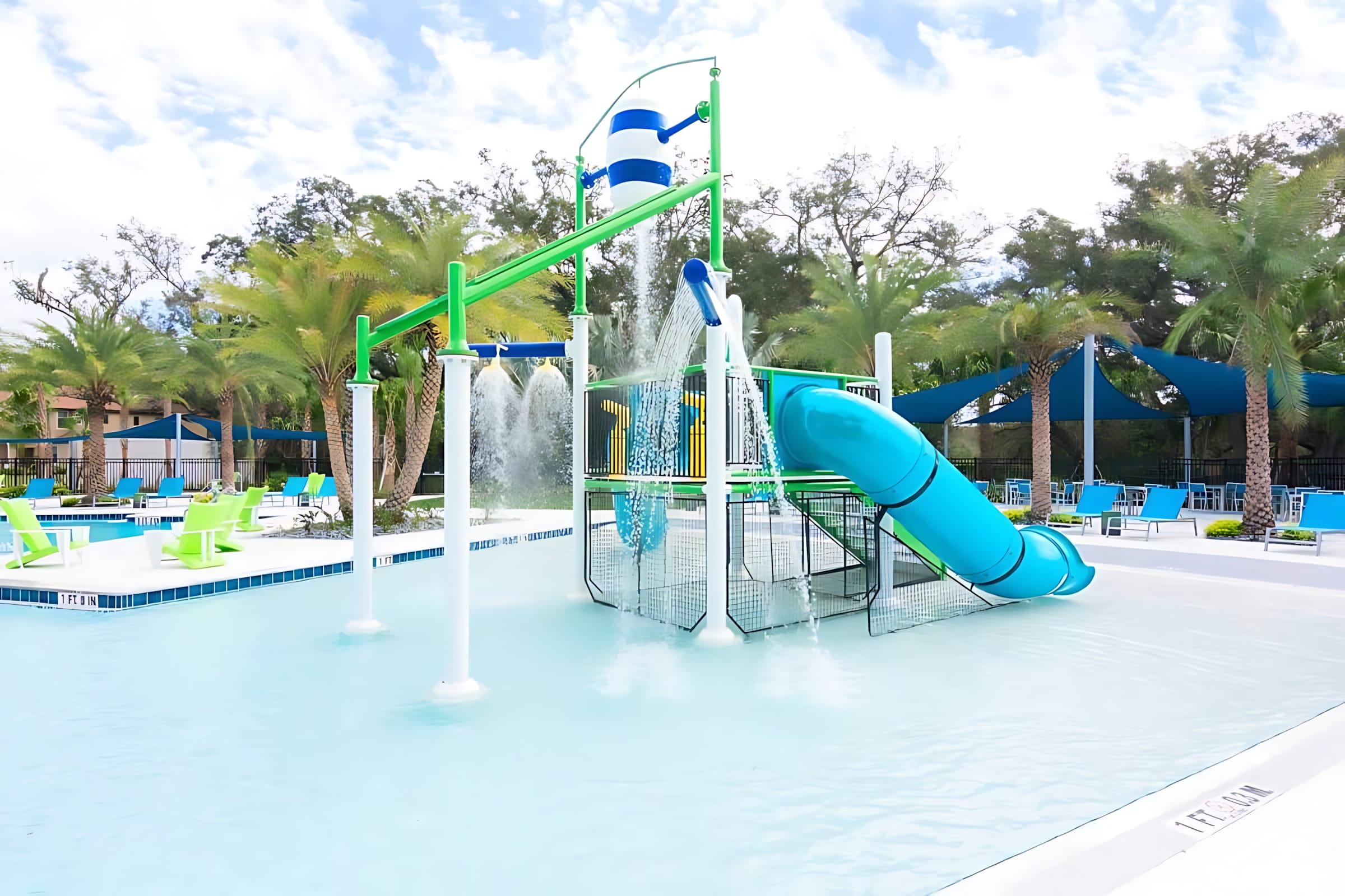 Kids' water play area within a communal pool, featuring a blue water slide, a tipping bucket, and various spray features. Palm trees and lounge chairs are visible in the background.
