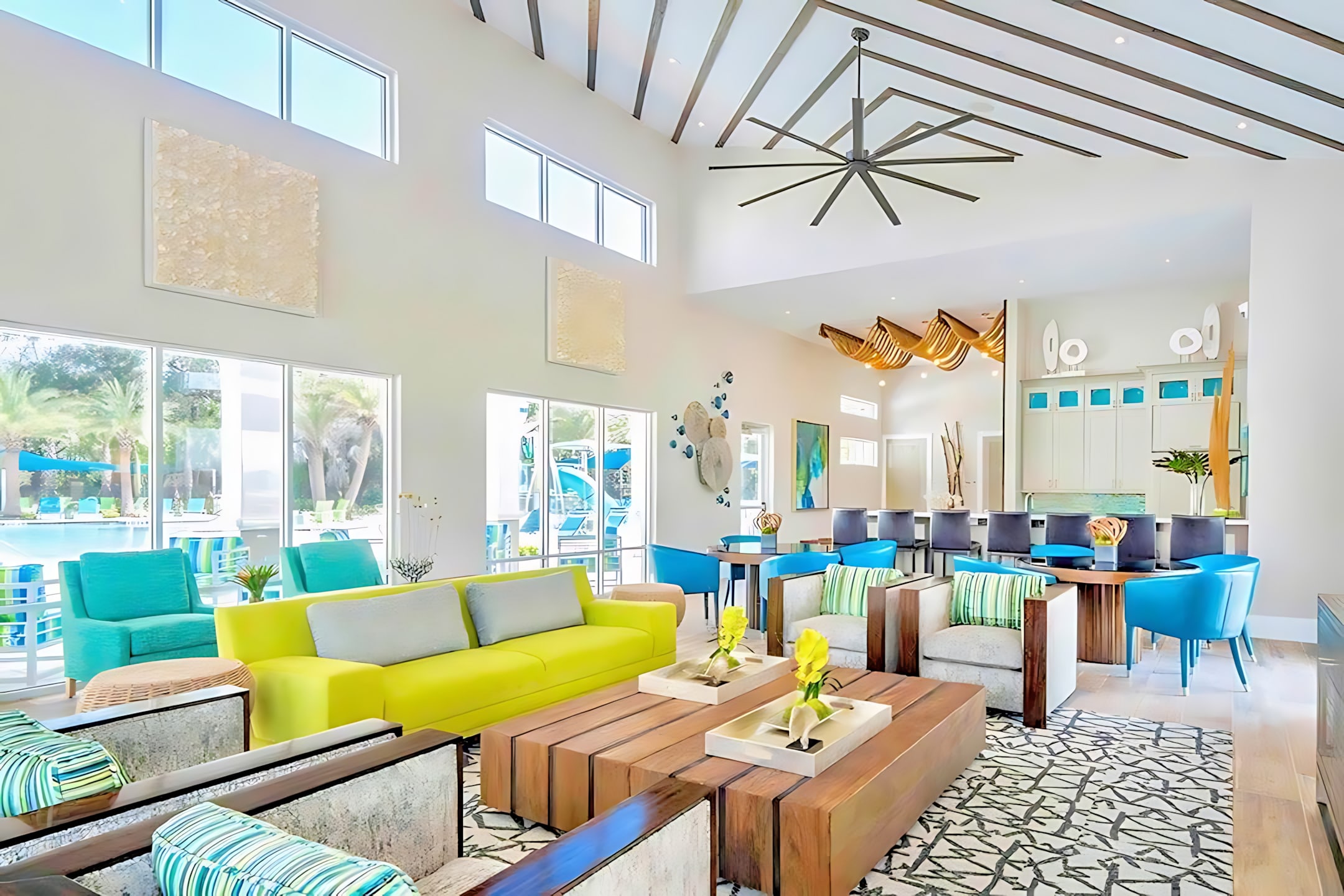 A bright and modern communal living area features a vibrant lime green sofa and two large wooden coffee tables on a patterned rug. High ceilings with exposed beams, large windows, and contemporary artwork create an airy, stylish atmosphere.