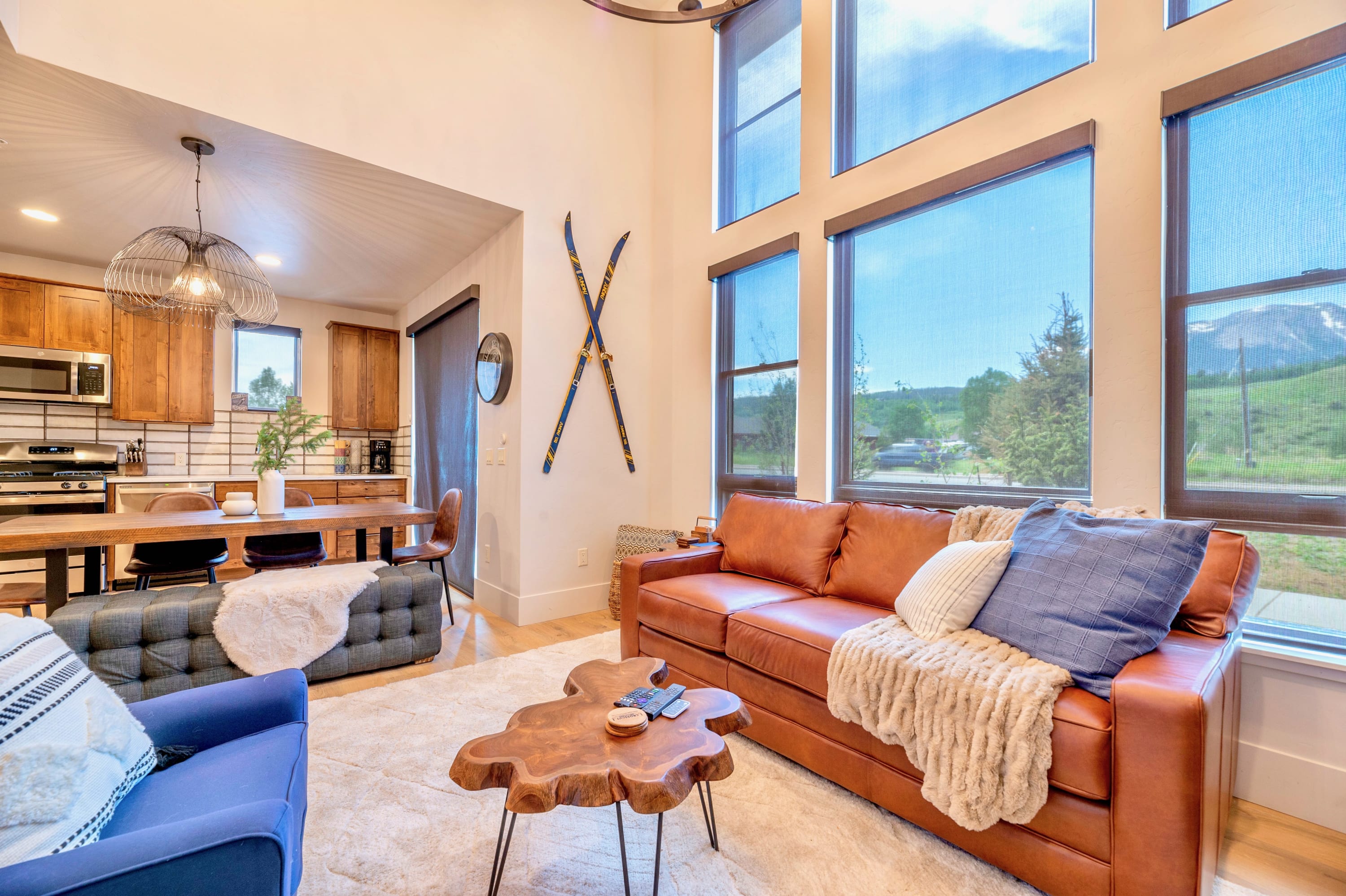 A spacious living room with a brown leather sofa and blue armchair on a light rug, featuring tall windows with mountain views and decorative skis mounted on the wall. The open-concept space flows into the kitchen and dining area.