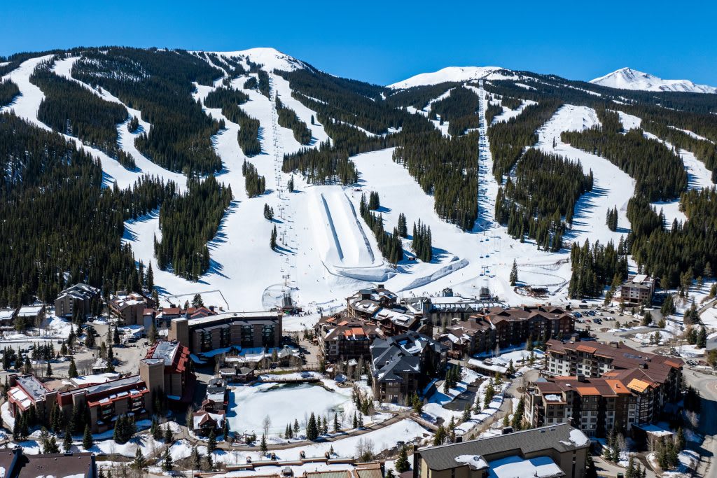 An aerial view captures a sprawling ski resort with snow-covered slopes, chairlifts, and a village nestled at the base, featuring numerous buildings with brown roofs and a partially frozen pond.