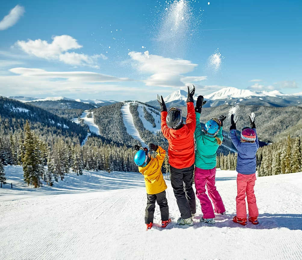 Four people, including children, in colorful winter clothing joyfully throw snow into the air on a sunny, snow-covered mountain slope. A vast ski resort with groomed runs and pine forests stretches into the background under a bright blue sky.