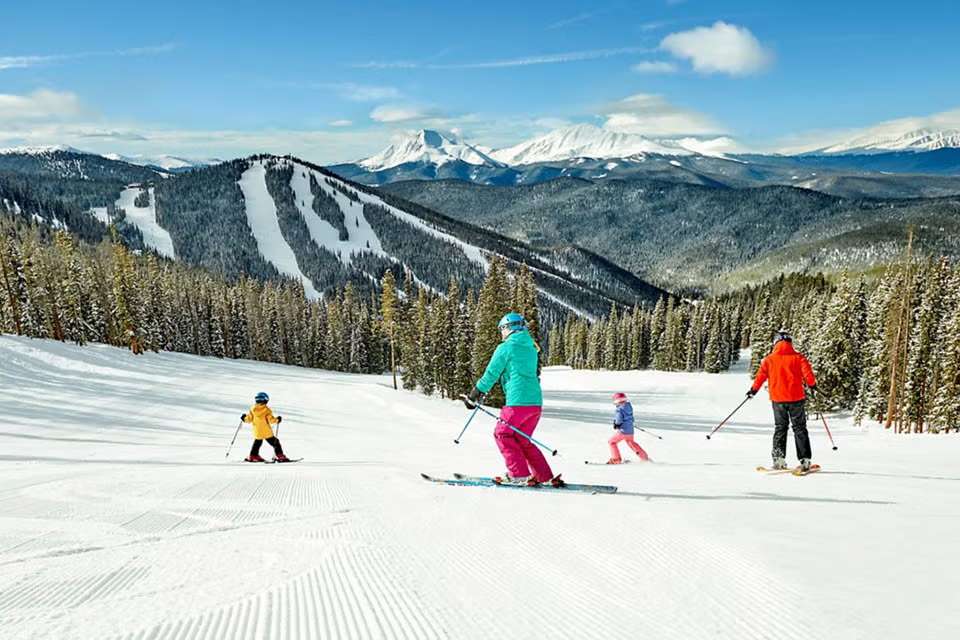 A family skis down a wide, groomed slope at a sunny mountain resort, with vast snow-covered peaks and pine forests in the background. The sky is bright blue with scattered clouds.