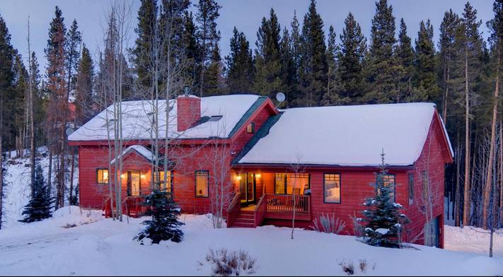 A charming red house with a snow-covered roof and warm interior lights glows amidst a dense pine forest, surrounded by deep snow. The winter scene is set against a soft twilight sky.