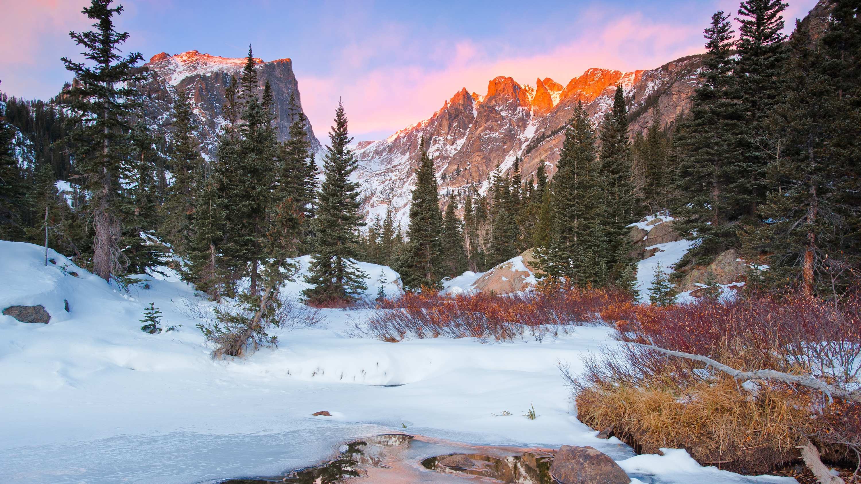 A stunning winter landscape showcases snow-covered mountains with peaks illuminated by a warm sunset glow, surrounded by dense evergreen forests and a partially frozen stream in the foreground.
