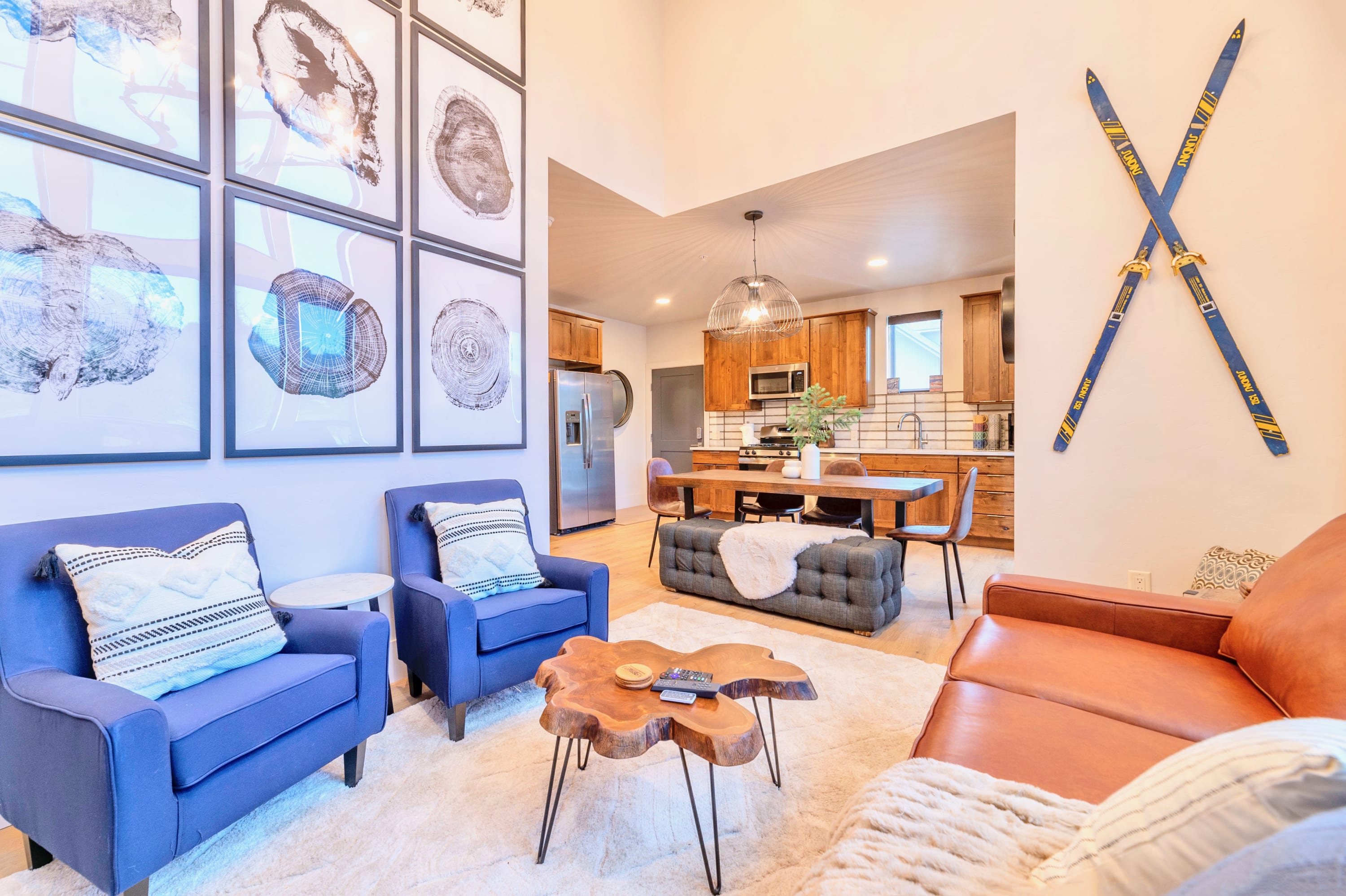 A bright living room with a brown leather sofa and two blue armchairs on a light rug, featuring a gallery wall of tree ring prints and a view into the adjacent kitchen and dining area. Decorative skis are mounted on the wall.