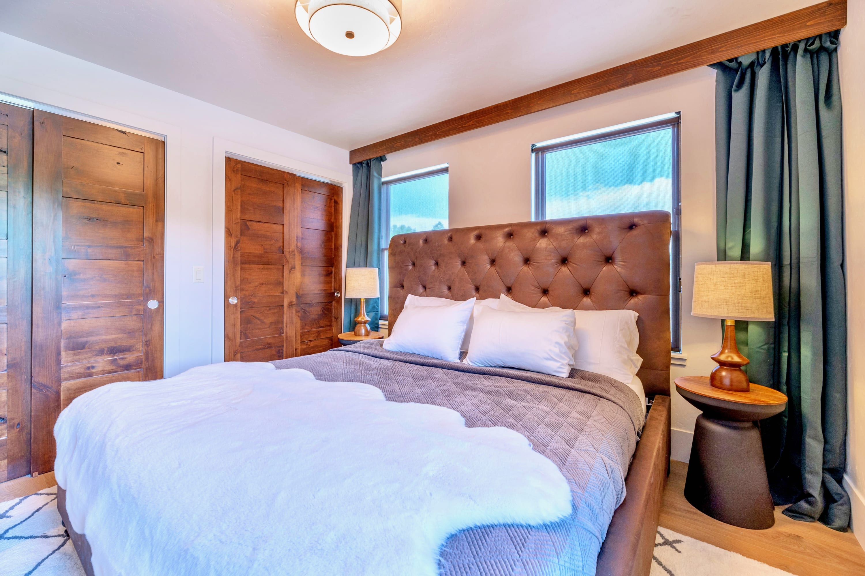 Cozy bedroom featuring a large bed with a brown tufted leather headboard, two rustic wooden closets, and small windows framed by dark teal curtains, creating a warm and inviting atmosphere.