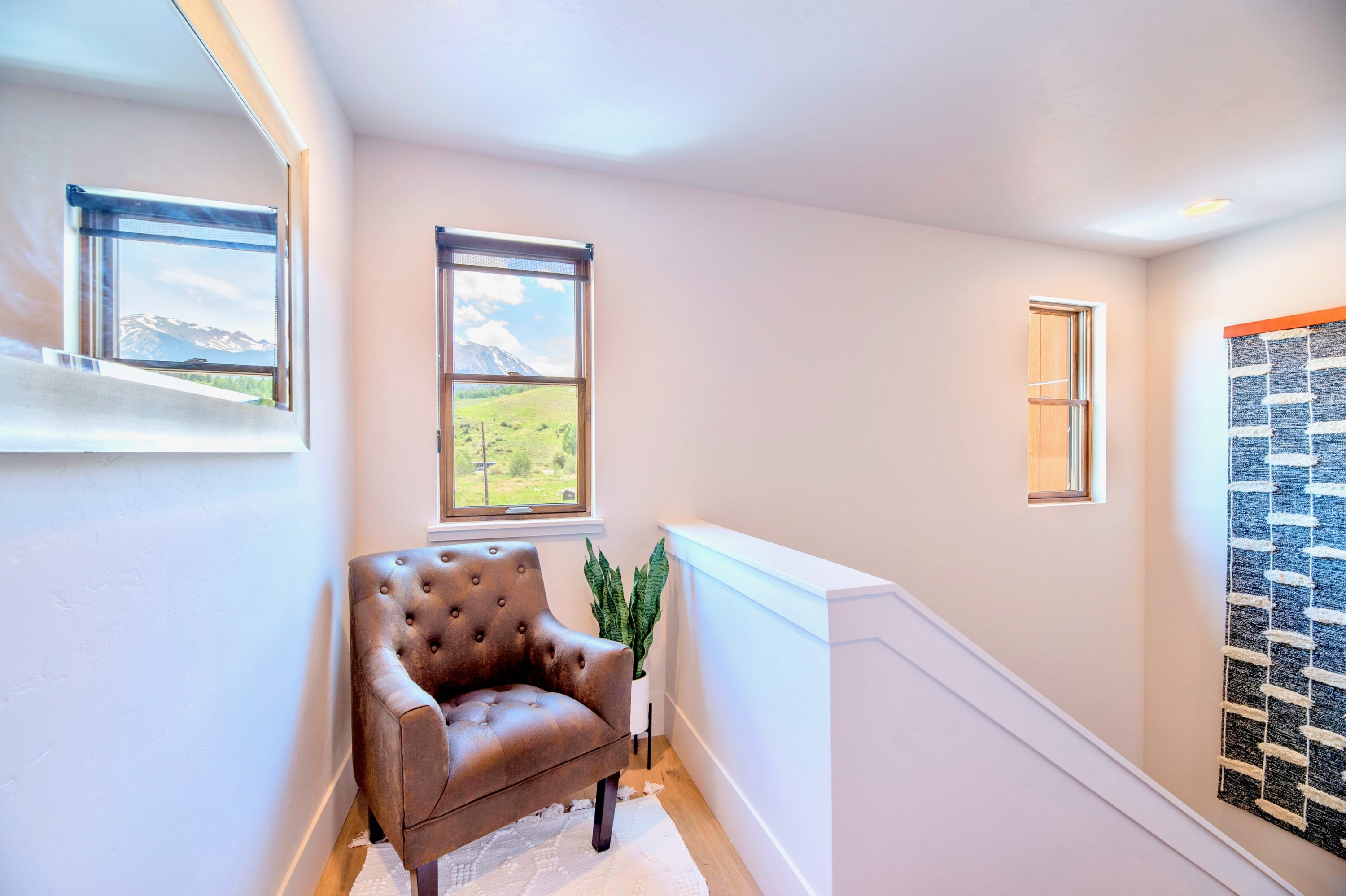 A bright hallway landing features a brown leather armchair, a snake plant, and a large window offering panoramic views of snow-capped mountains and green hills. A mirror reflects the outdoor scenery, and a textured wall hanging adds a decorative touch.
