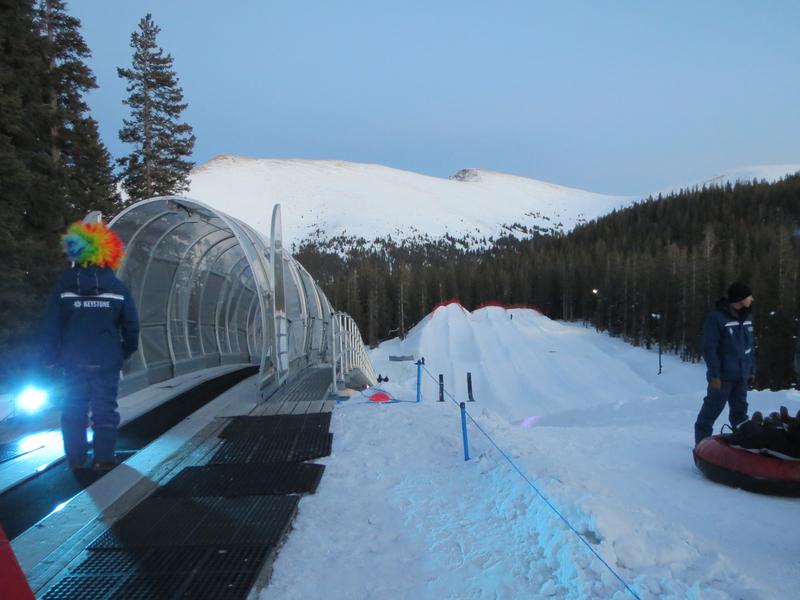 A snow tubing hill at dusk features a covered conveyor lift and several snow lanes, with people in winter gear and a person in a colorful wig. Snow-covered mountains and pine trees are visible in the background under a soft blue sky.