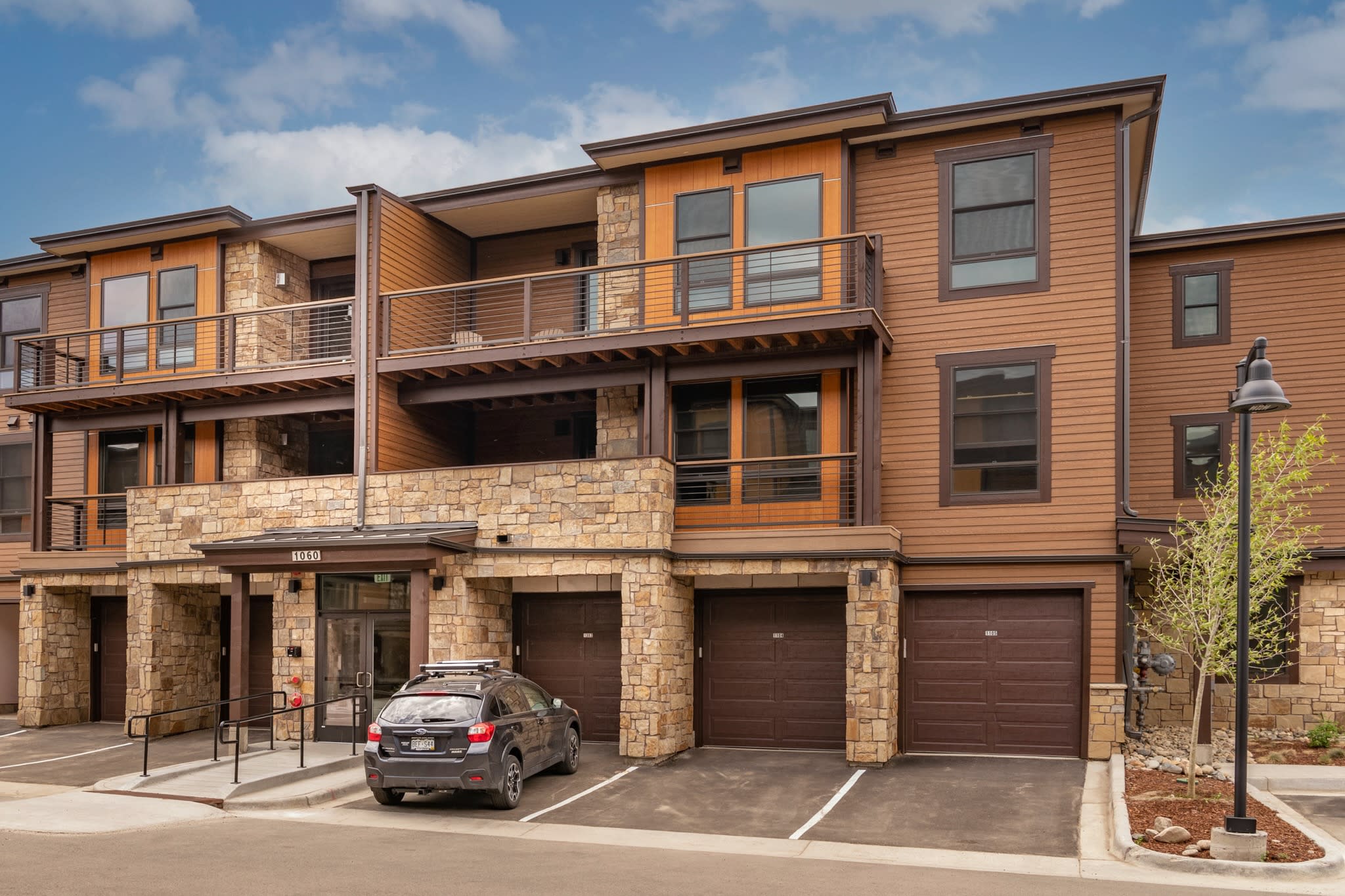 A modern multi-story townhouse building features a facade of brown siding and natural stone, with multiple balconies and attached garages. A dark SUV is parked in front of the entrance, under a partly cloudy sky.