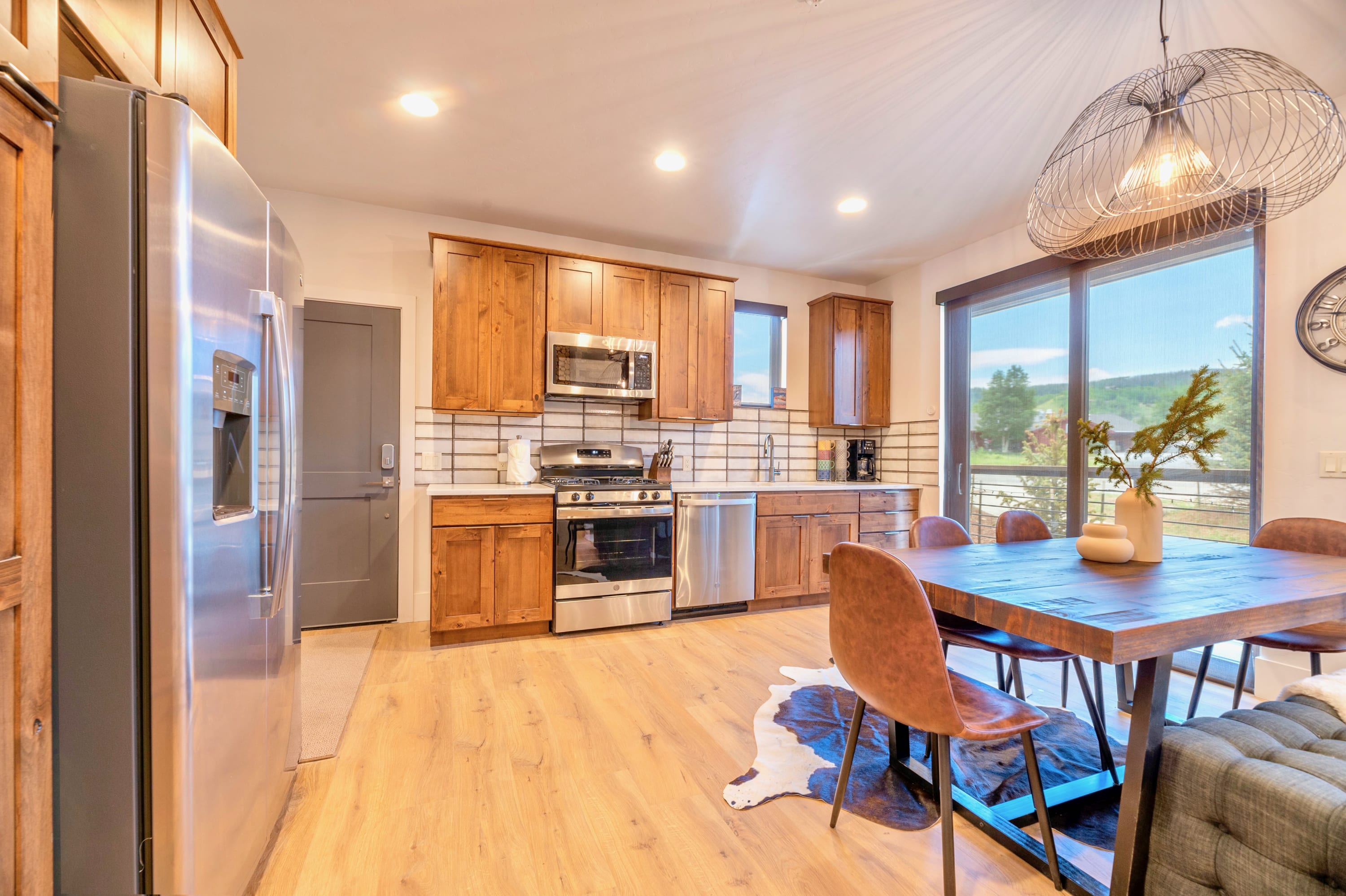 Bright open-concept kitchen and dining space with light hardwood floors, featuring wooden cabinetry, stainless steel appliances, a rustic dining table, and large windows offering mountain views.