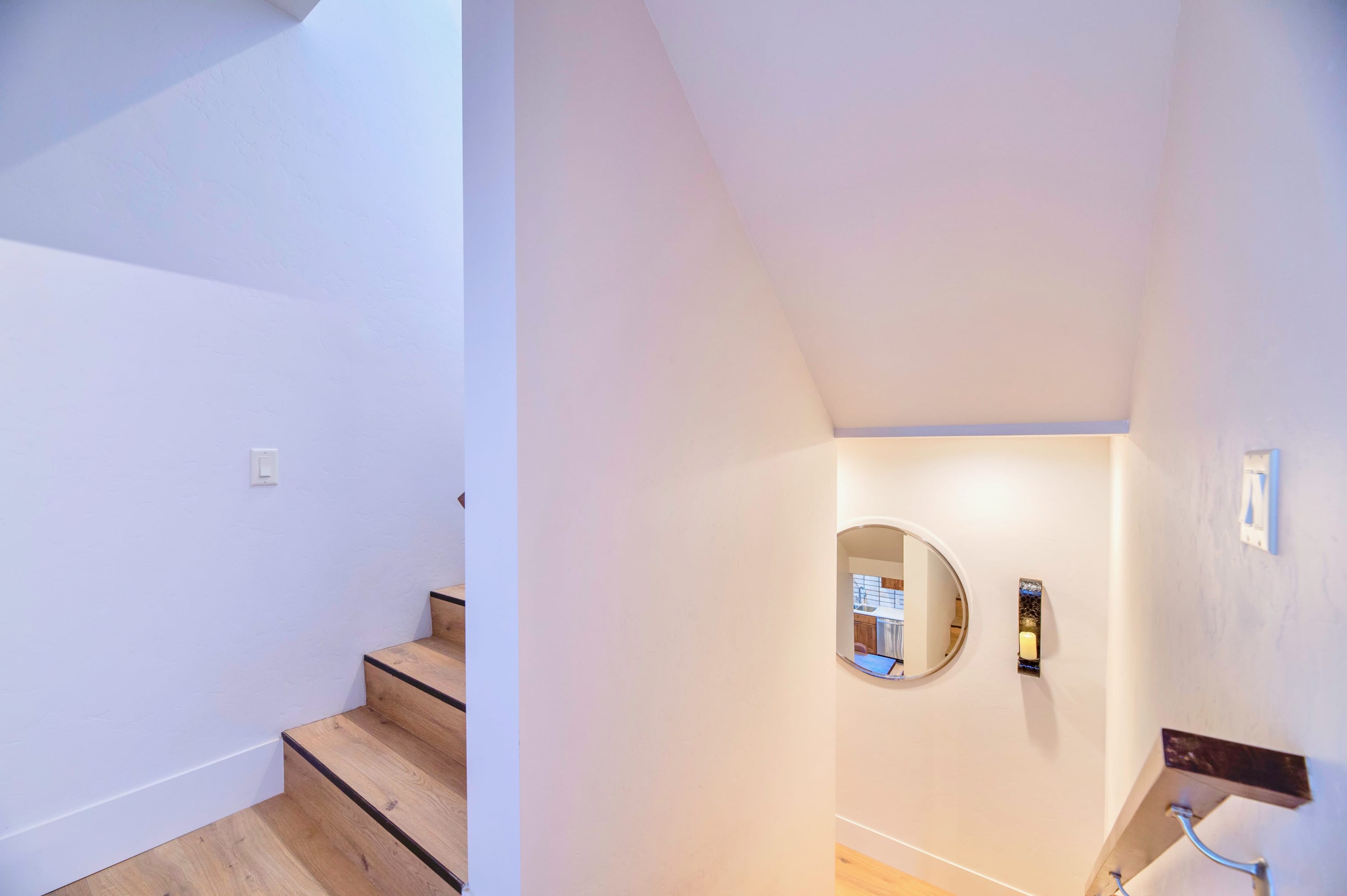 Well-lit interior staircase with light wooden treads and dark risers, featuring white walls and a small round mirror on the landing, leading to another level of the home.