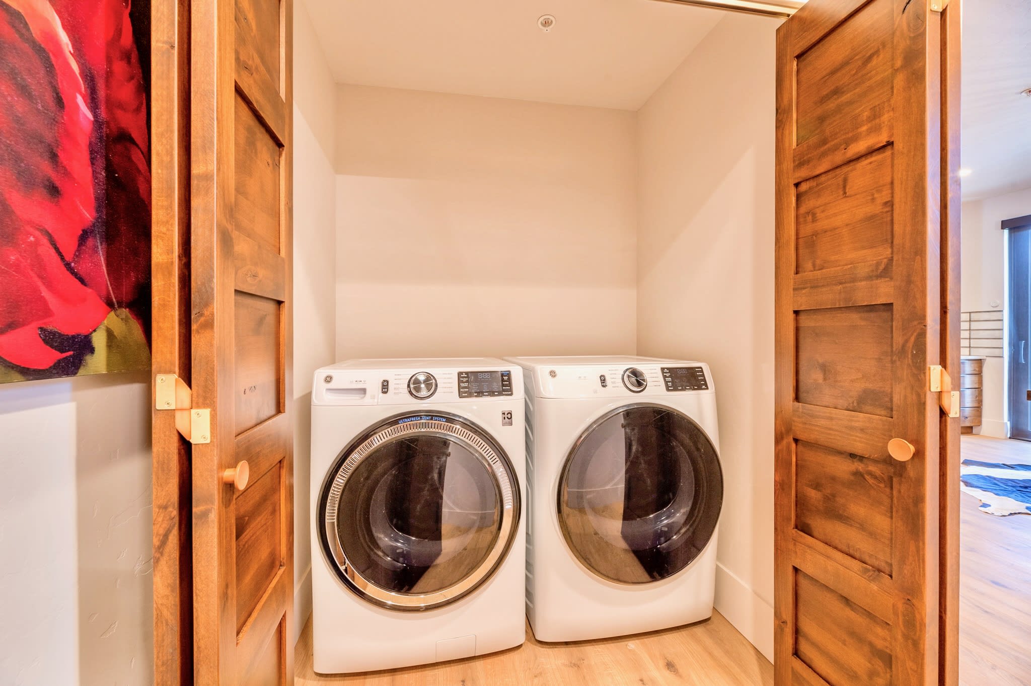 A compact laundry closet features a white front-loading washer and dryer set, neatly tucked behind open wooden bi-fold doors. The space has light-colored walls and light wood flooring.
