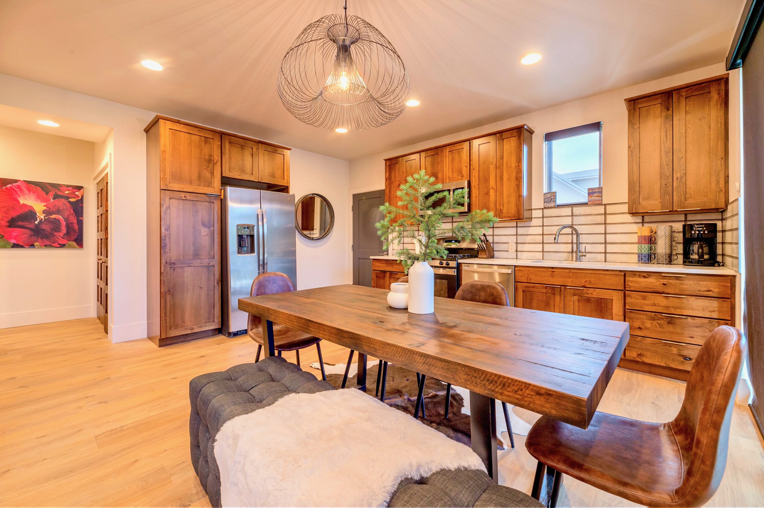 Modern dining area featuring a large rustic wooden table with a dark upholstered bench and leather chairs, illuminated by a unique wire-frame chandelier, with a full kitchen visible in the background.