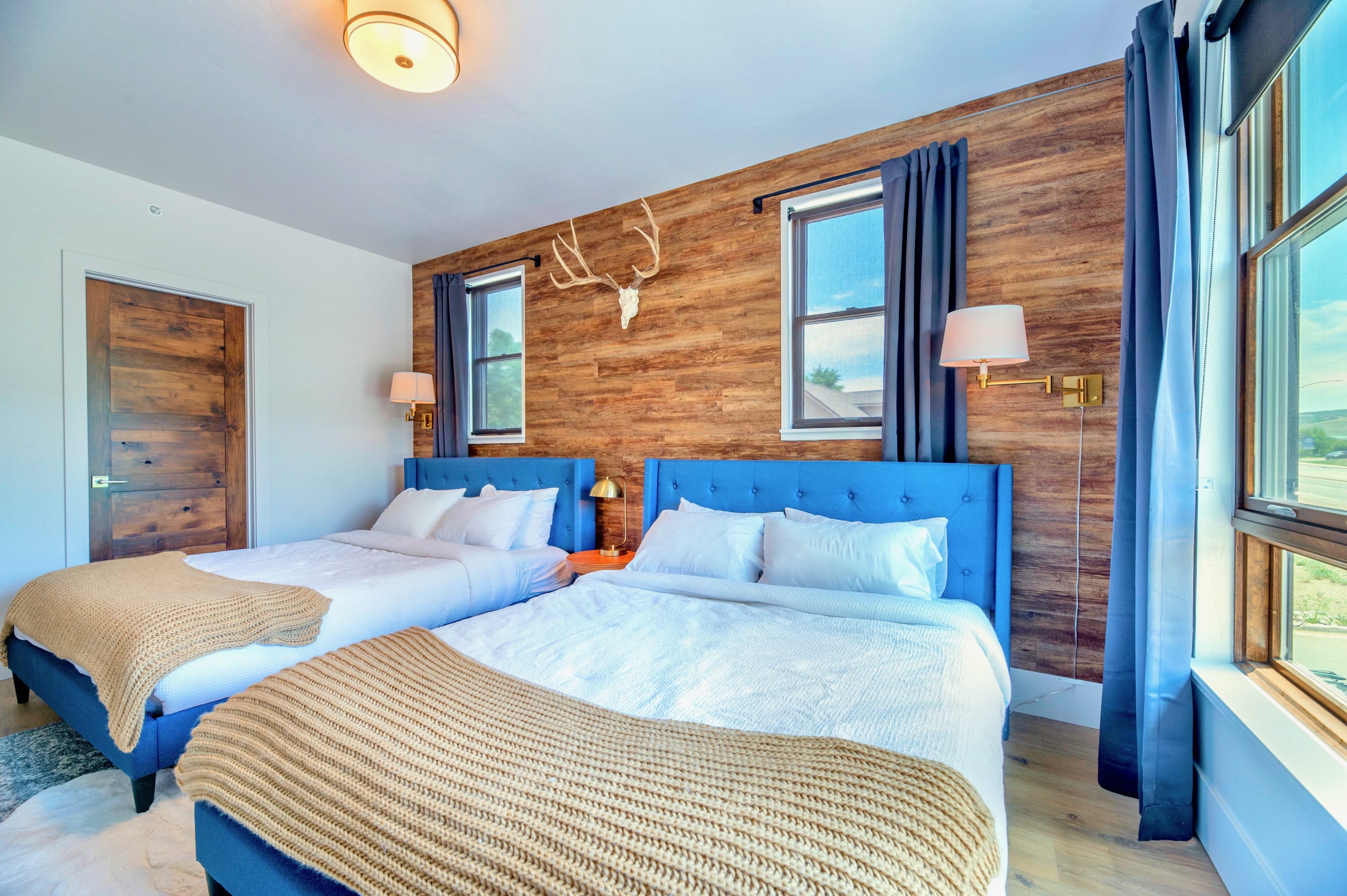 Another view of the second bedroom, highlighting the two beds with blue upholstered headboards, the striking wood plank accent wall with antler decor, and bright windows.