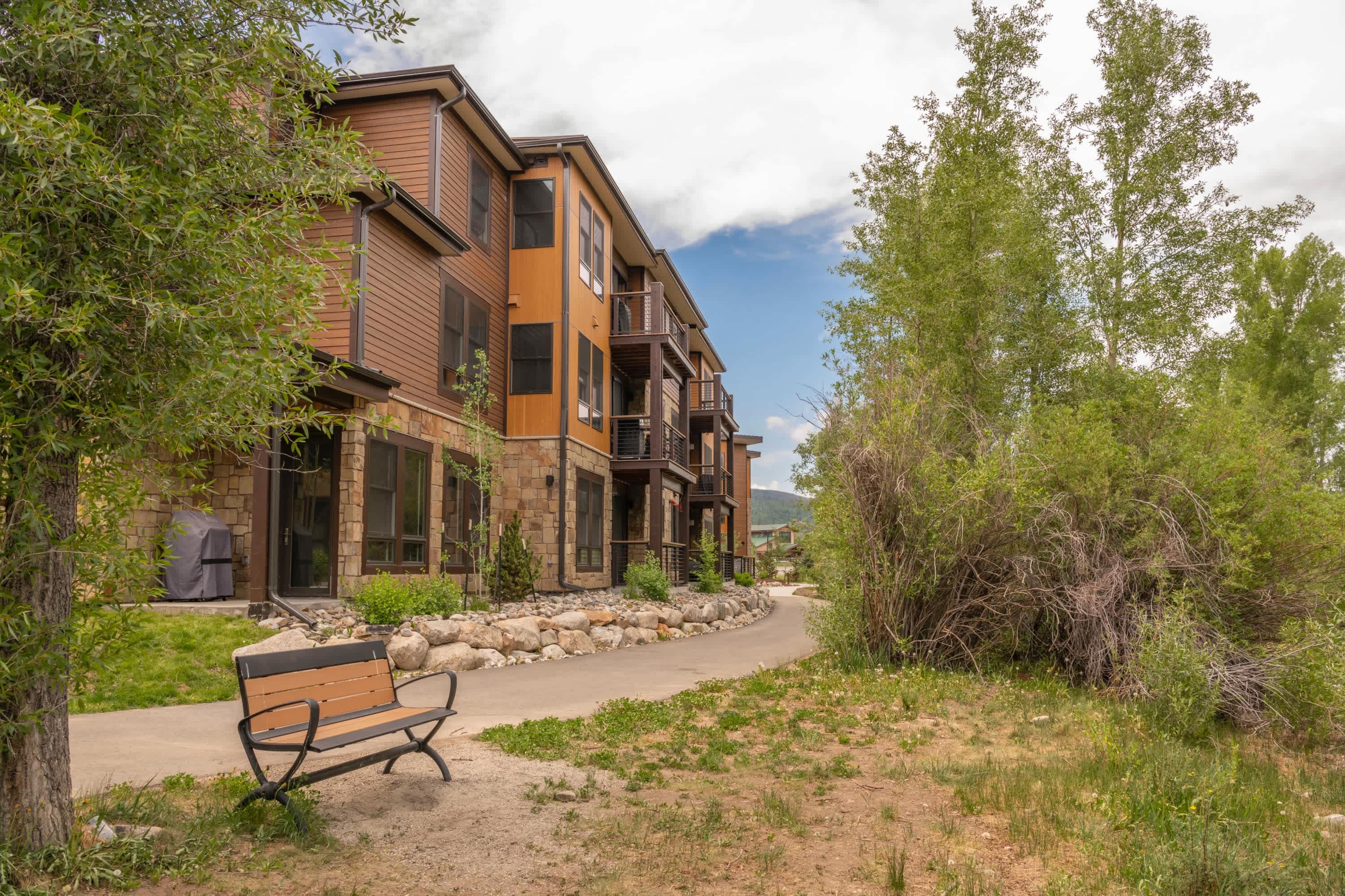 The side of a modern townhouse building, constructed with brown siding and stone, is visible alongside a paved walking path and lush green landscaping. A wooden bench sits beside the path, surrounded by trees and shrubs under a cloudy sky.