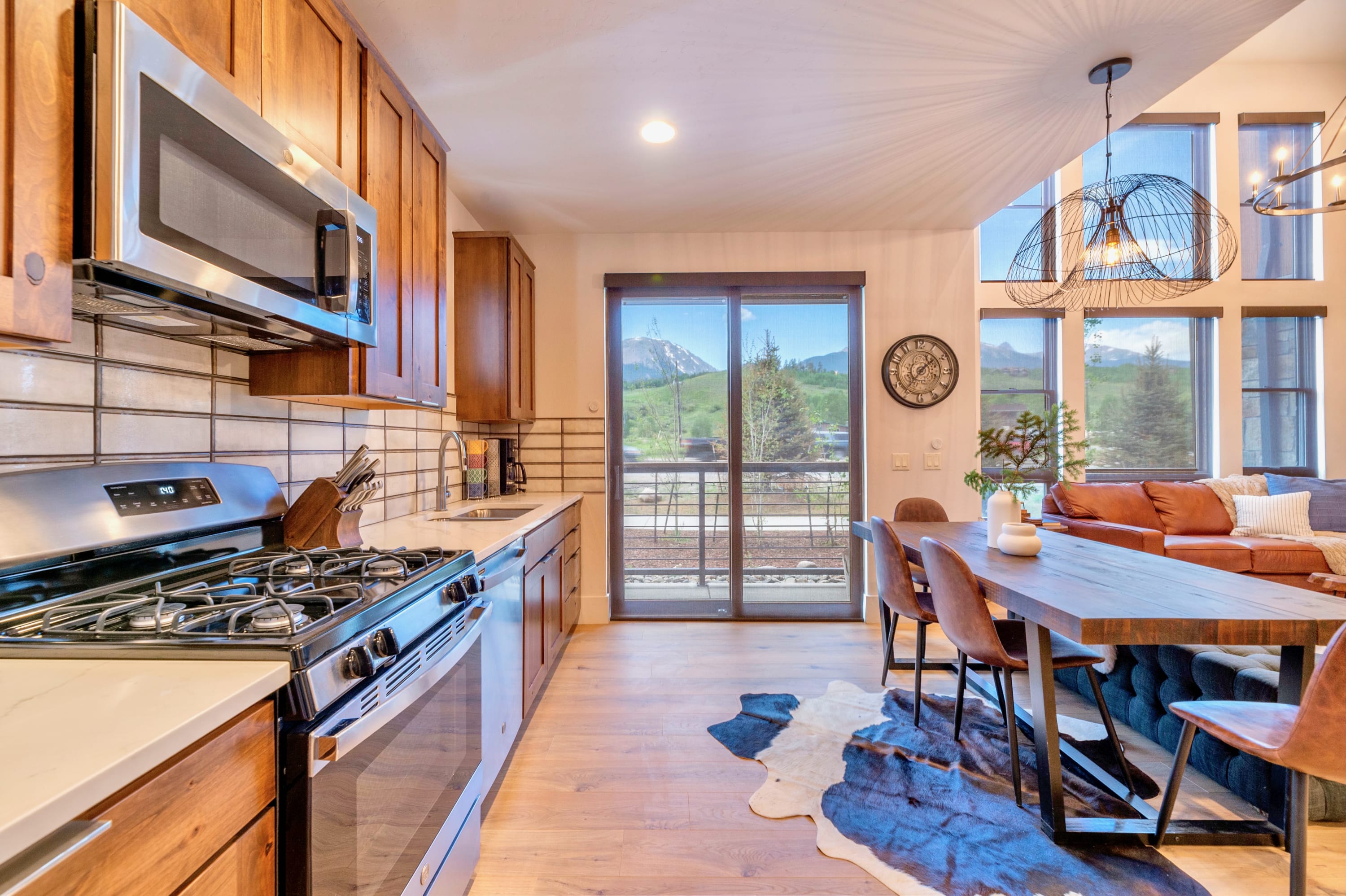 Well-equipped kitchen with light wooden cabinets, white subway tile backsplash, stainless steel appliances including a gas range, and a large window overlooking scenic mountain landscapes.
