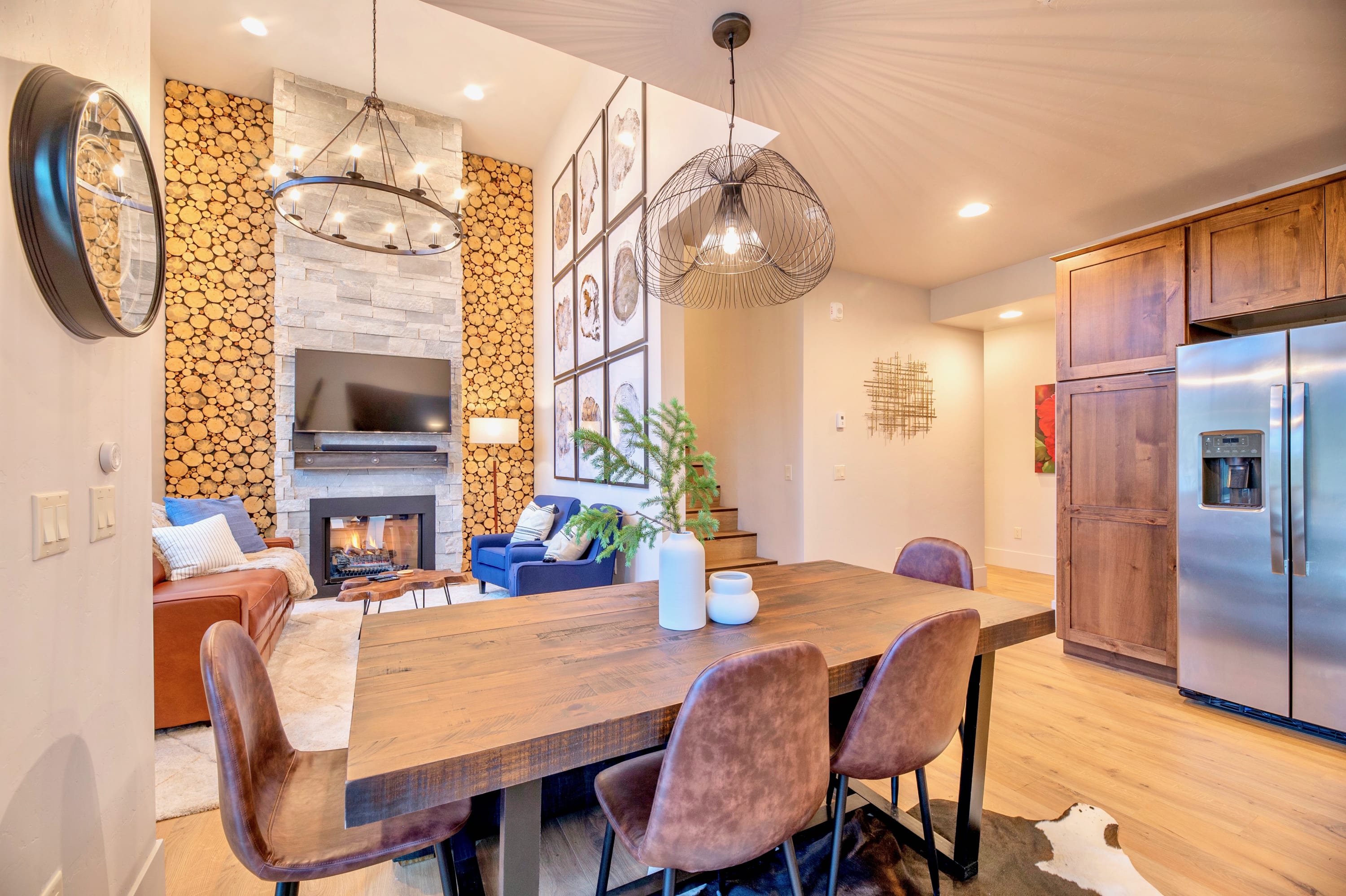 A rustic wooden dining table with four brown chairs and a bench sits on light wood flooring, with a view of the living room's stone fireplace and log accent wall. The kitchen with stainless steel appliances is visible to the right.