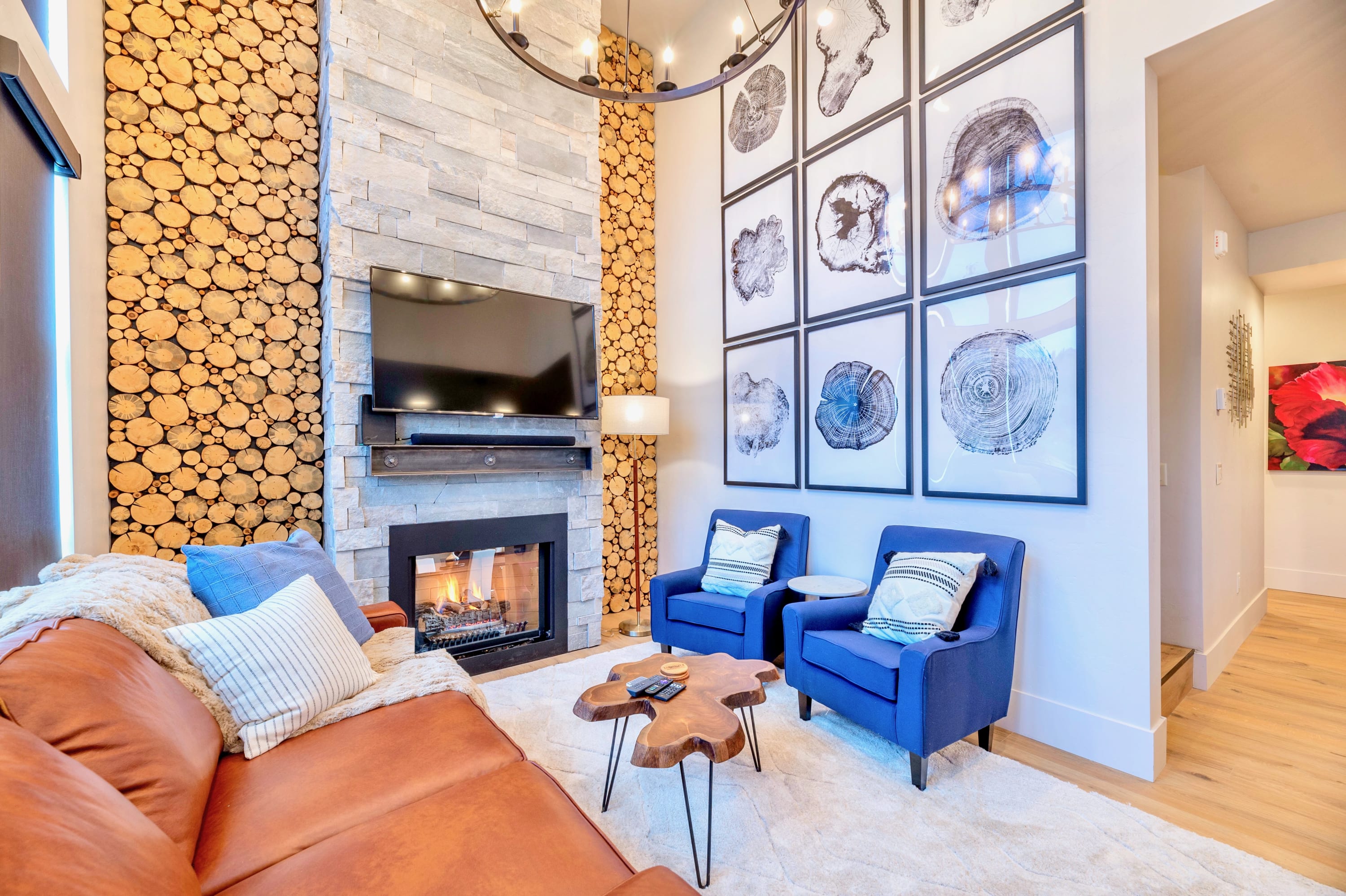A cozy living room features a brown leather sofa and two blue armchairs on a light rug, facing a stone fireplace with a mounted TV and decorative log paneling. A gallery wall with framed tree ring prints adorns the adjacent wall.