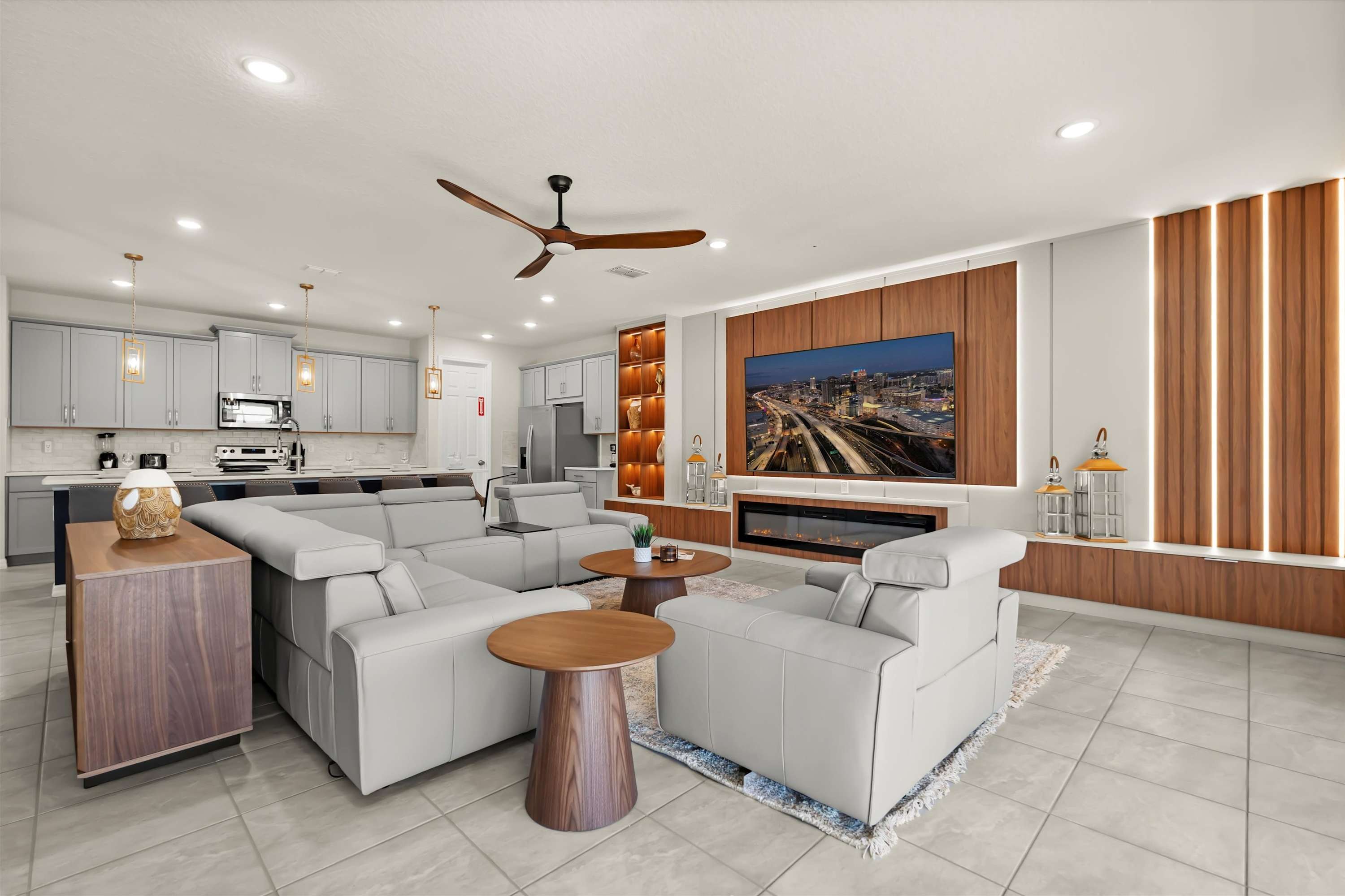 Wide view of a modern open-concept living room with a light gray sectional sofa, two armchairs, and a large wall-mounted TV above a fireplace, seamlessly flowing into a contemporary kitchen with gray cabinets and a breakfast bar.