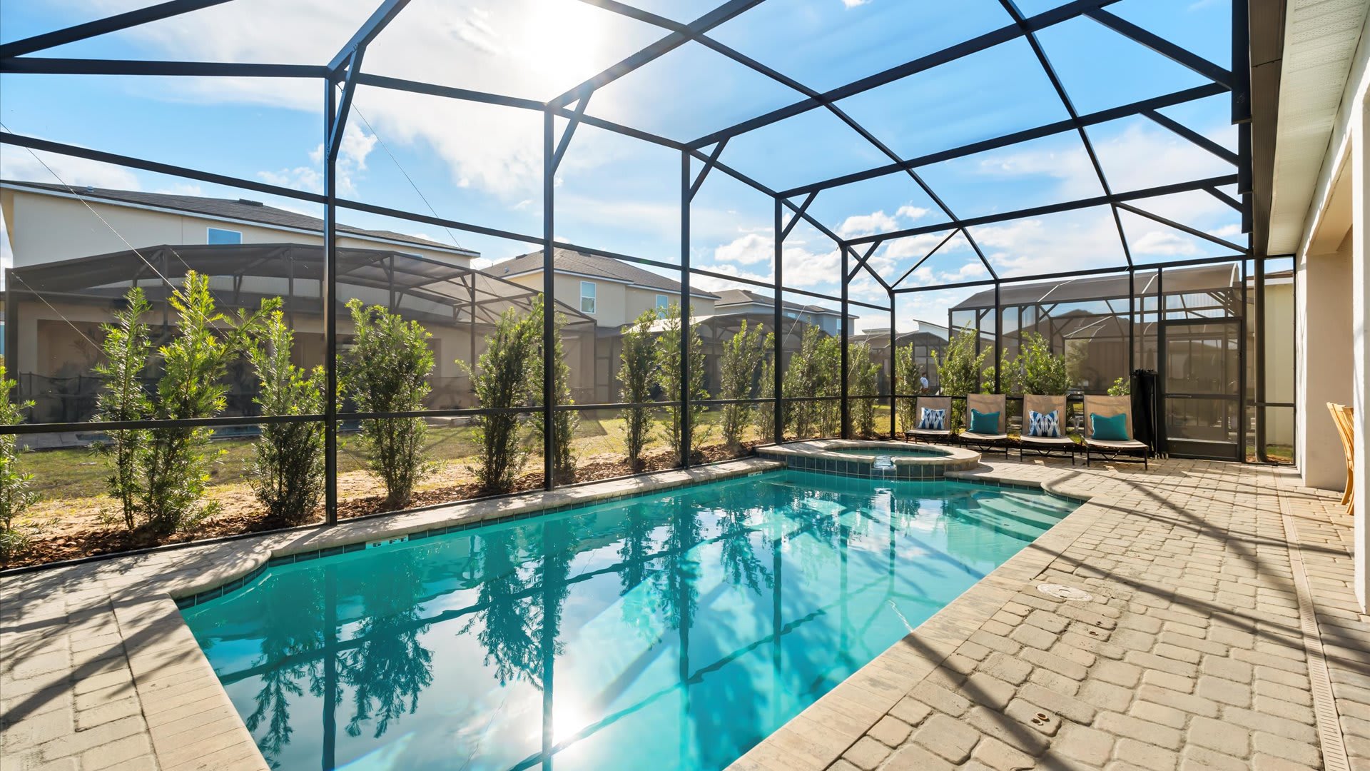 Bright outdoor private pool area enclosed by a screen, featuring a rectangular pool, a hot tub, and lounge chairs, with small trees lining the back fence and houses visible in the distance.