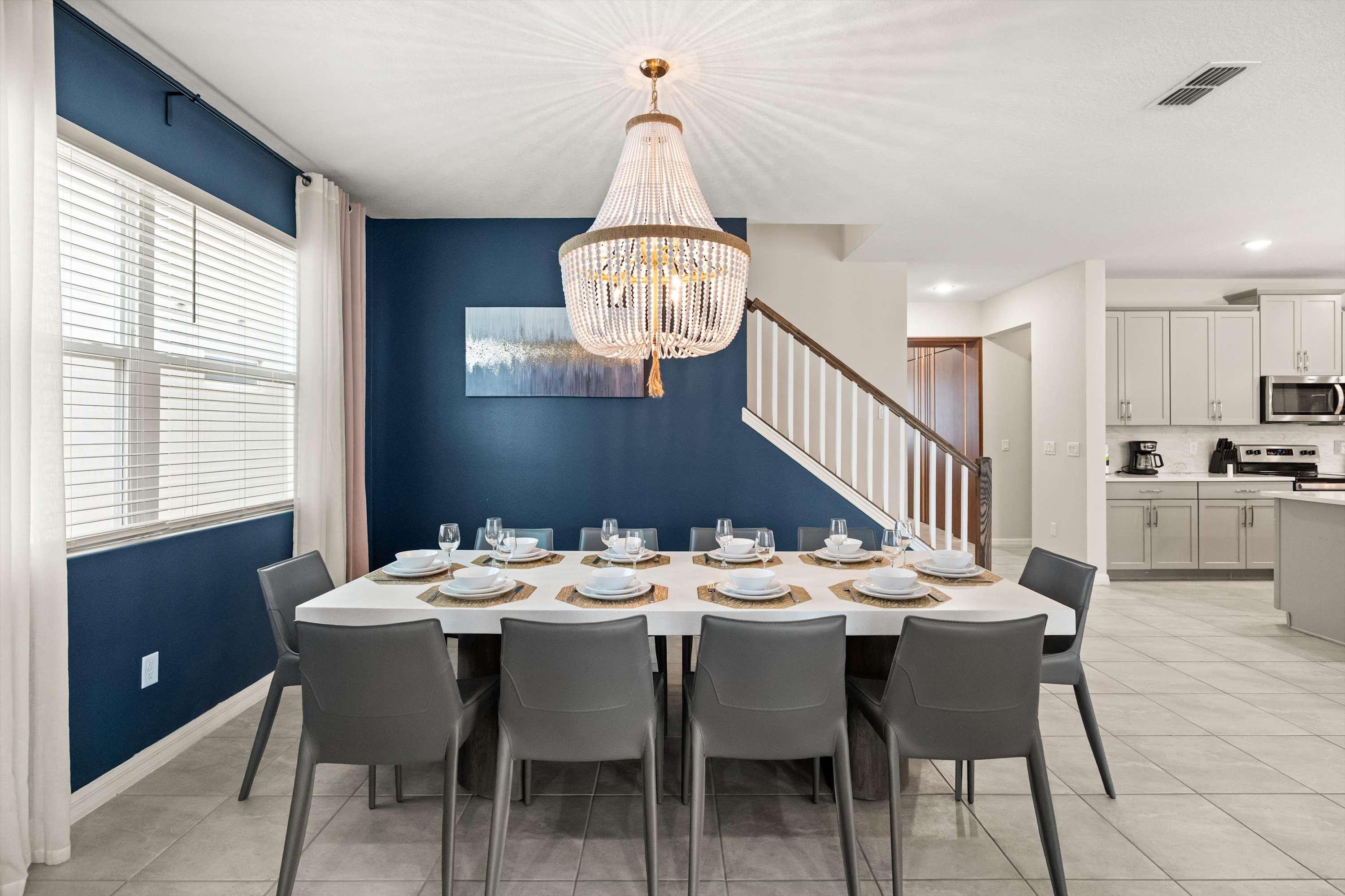 A spacious dining room features a long white table set for eight, surrounded by dark gray chairs, against a striking dark blue accent wall with modern art. A large, ornate crystal chandelier hangs overhead, and a staircase is visible in the background.