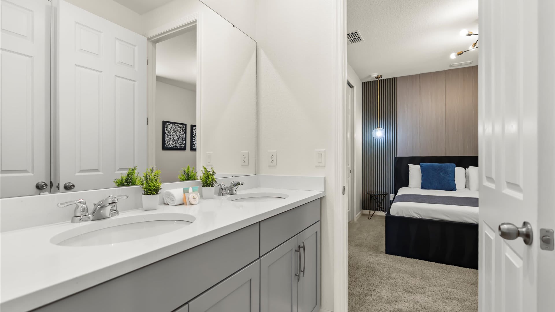 Double vanity bathroom with light gray cabinets, white countertops, and two sinks, seen from the doorway leading into bedroom-3. A large mirror reflects the bedroom, and small potted plants decorate the counter.