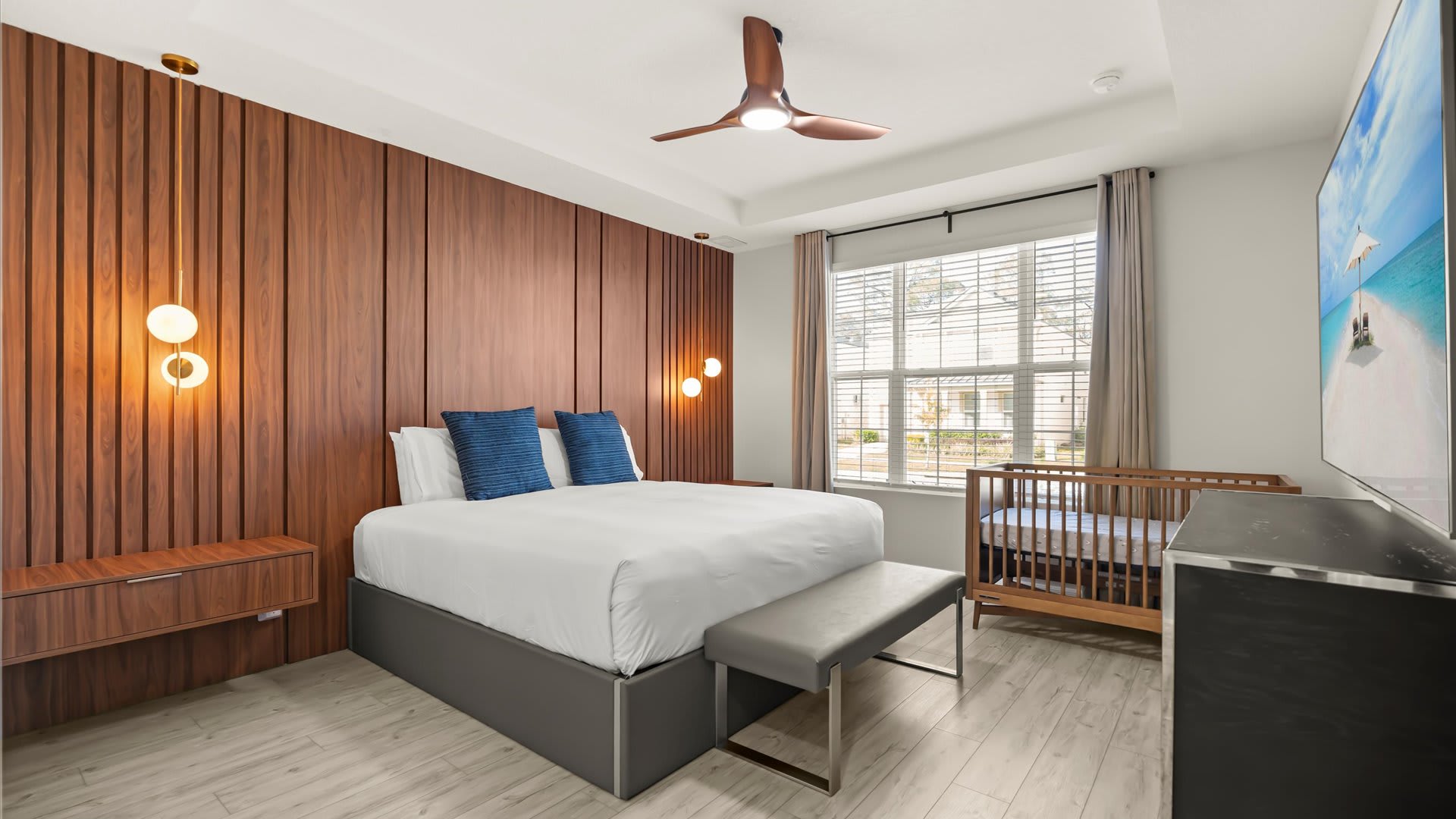 A modern bedroom features a king-sized bed with a grey frame and blue pillows, set against a striking dark wood slatted accent wall with integrated sconce lighting. A wooden crib is positioned near the window, and light wood-look flooring covers the room.
