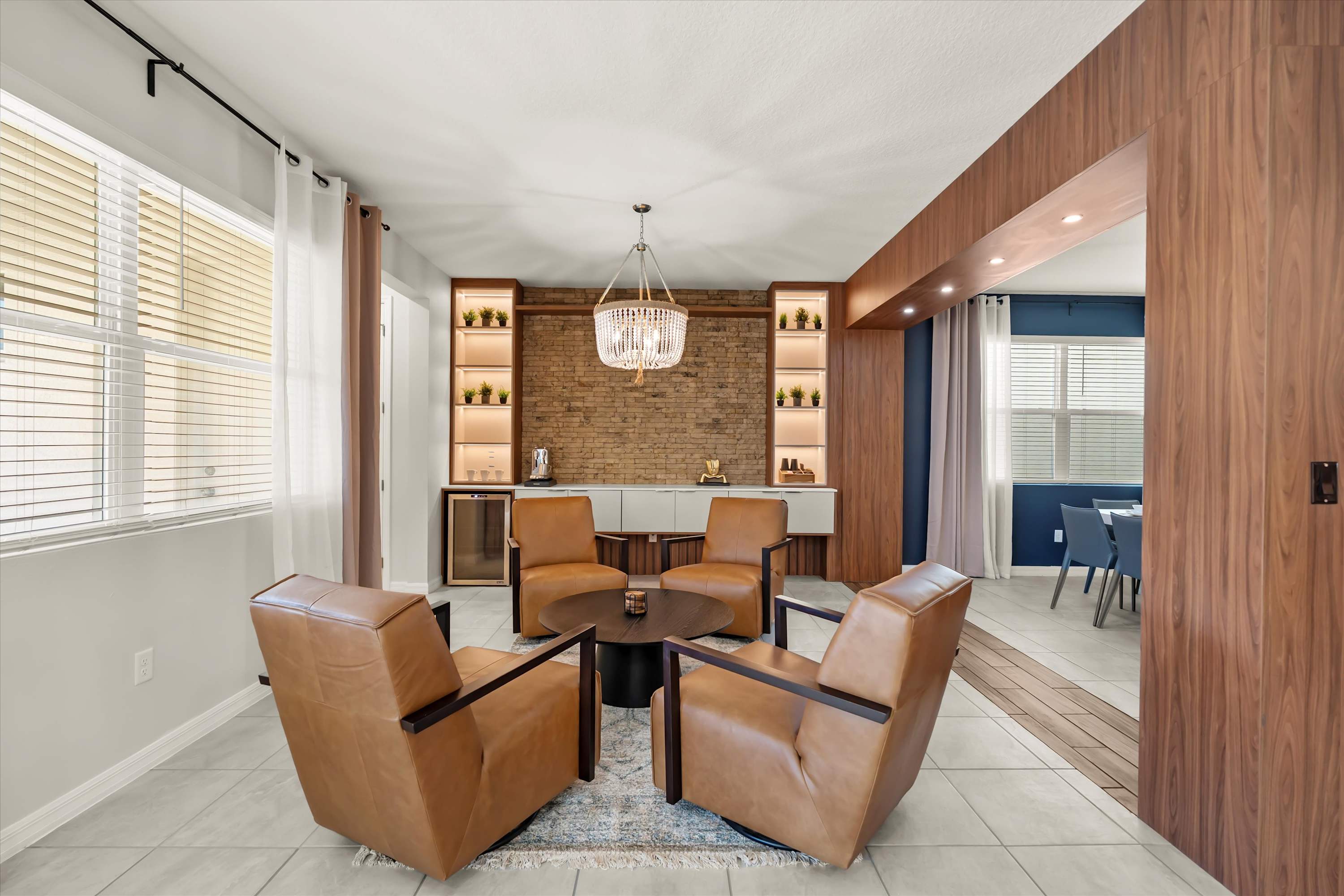 A cozy lounge area features four tan leather armchairs arranged around a dark round coffee table, set on a patterned rug. Behind them, a brick accent wall is flanked by illuminated built-in shelves and a beverage cooler, all beneath a crystal chandelier.