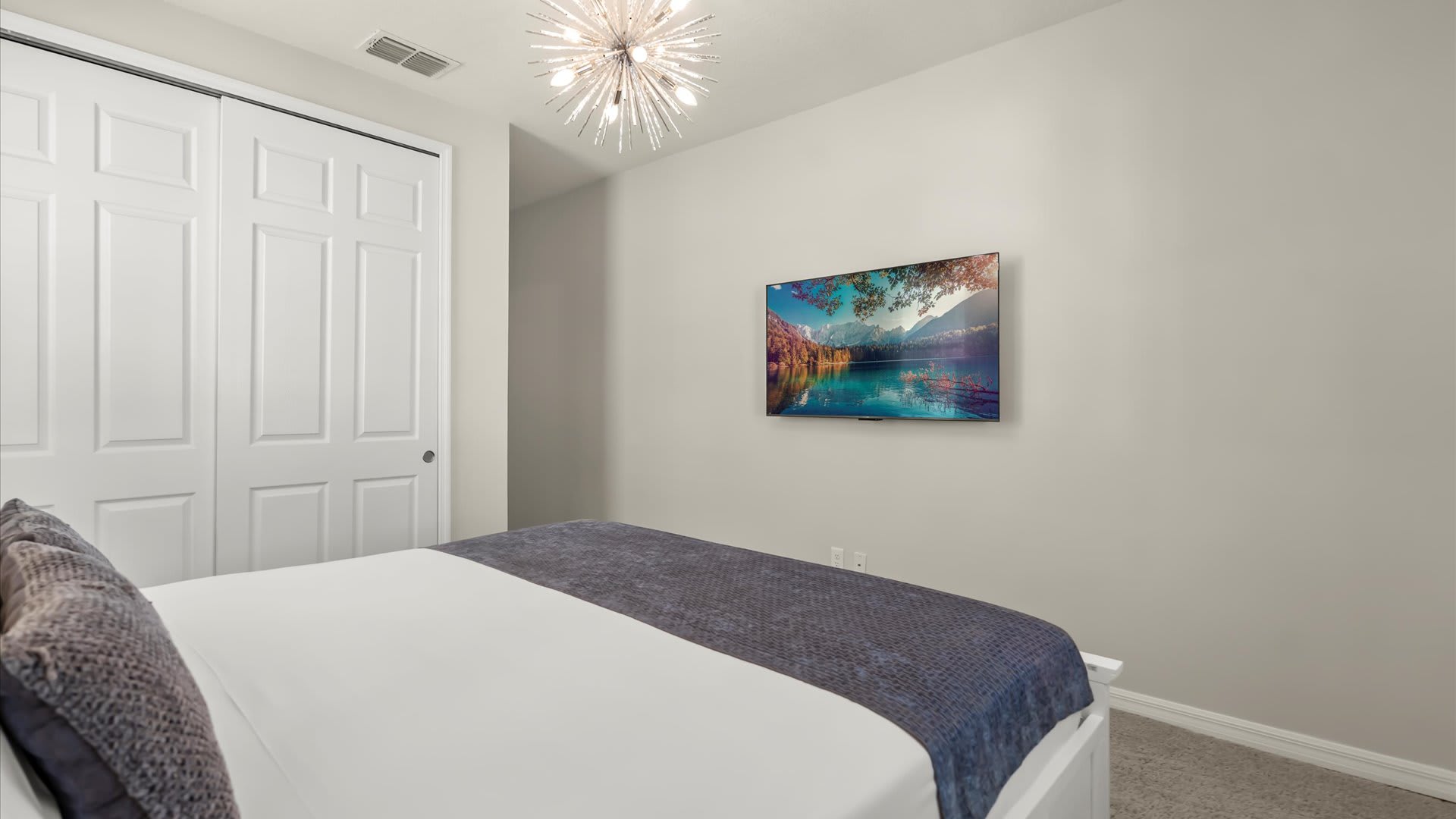 View of bedroom-2 showing a wall-mounted TV displaying a lake scene, a white bed with gray throw, and a white bi-fold closet door. The room has light gray walls and a sputnik-style chandelier.